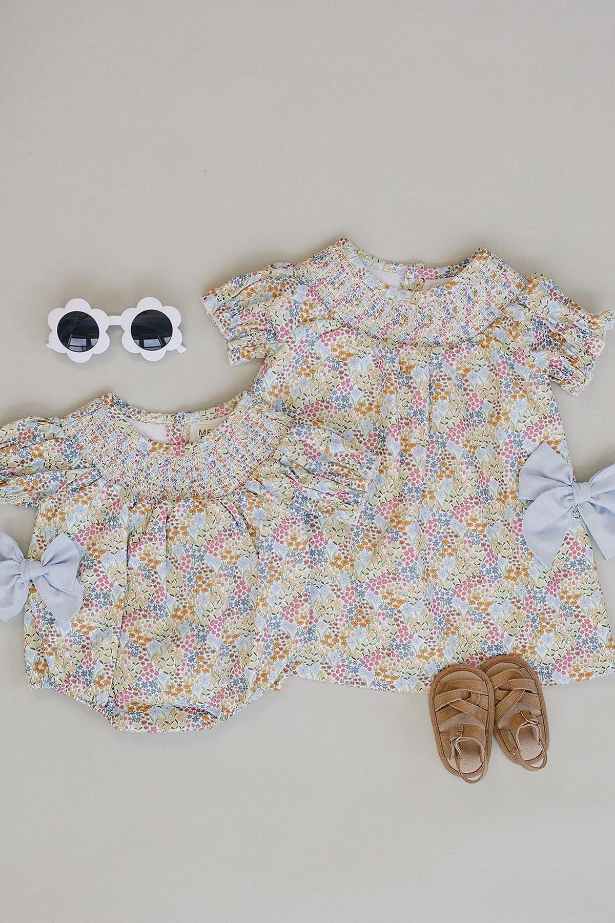 Two baby dresses in a cheerful floral pattern, featuring soft pastel colors and puffed sleeves, accompanied by a pair of brown sandals and white sunglasses, displayed on a neutral background.