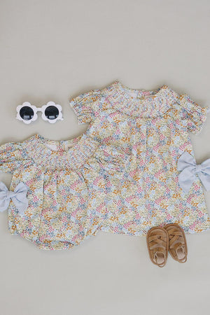 Two baby dresses in a cheerful floral pattern, featuring soft pastel colors and puffed sleeves, accompanied by a pair of brown sandals and white sunglasses, displayed on a neutral background.