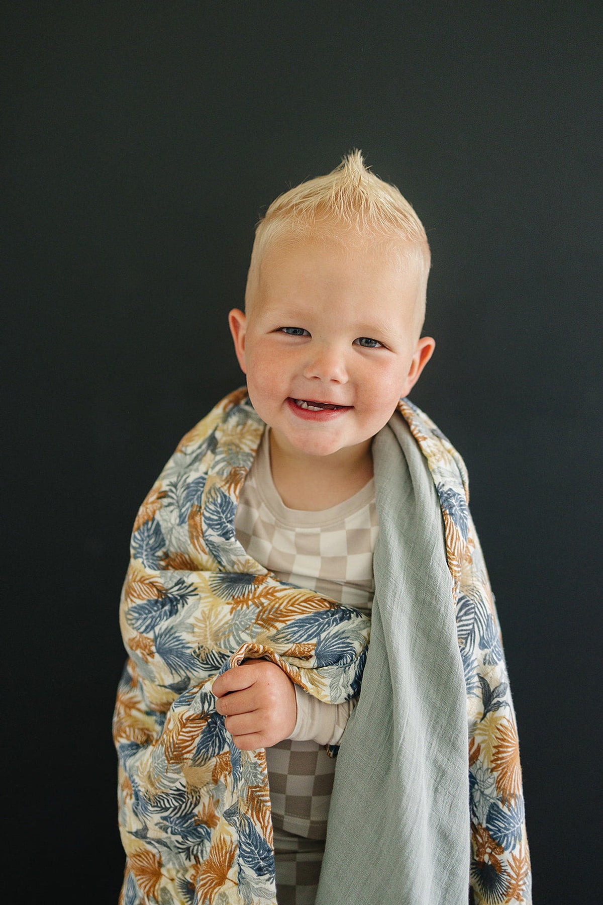 A smiling child wrapped in a colorful, leafy-patterned muslin quilt with a dark background.