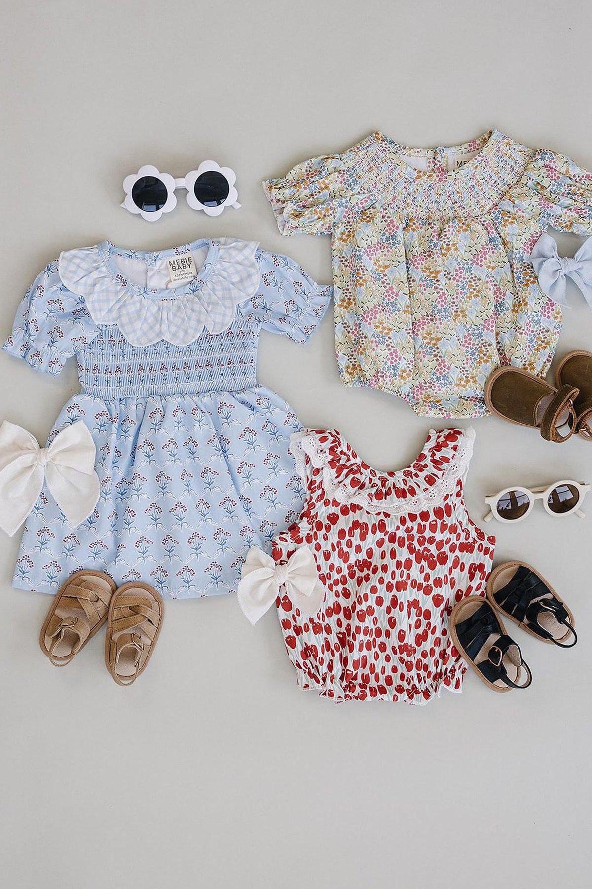 Three adorable baby dresses are displayed: a light blue dress with a gingham collar and red floral accents, a colorful floral print dress, and a red and white spotted romper.