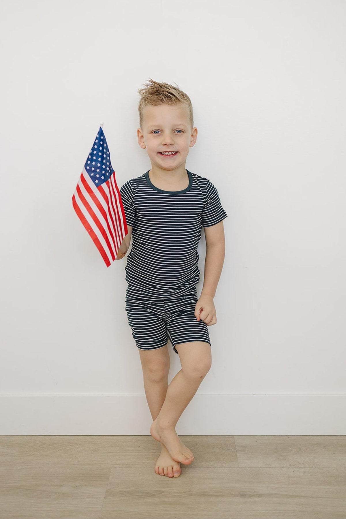 A child in navy striped shorts set holding a small American flag, standing against a white wall.