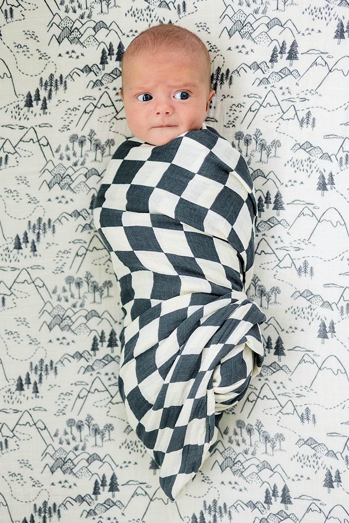 A baby is swaddled in a charcoal checkered muslin blanket, lying on a mountain-patterned background.
