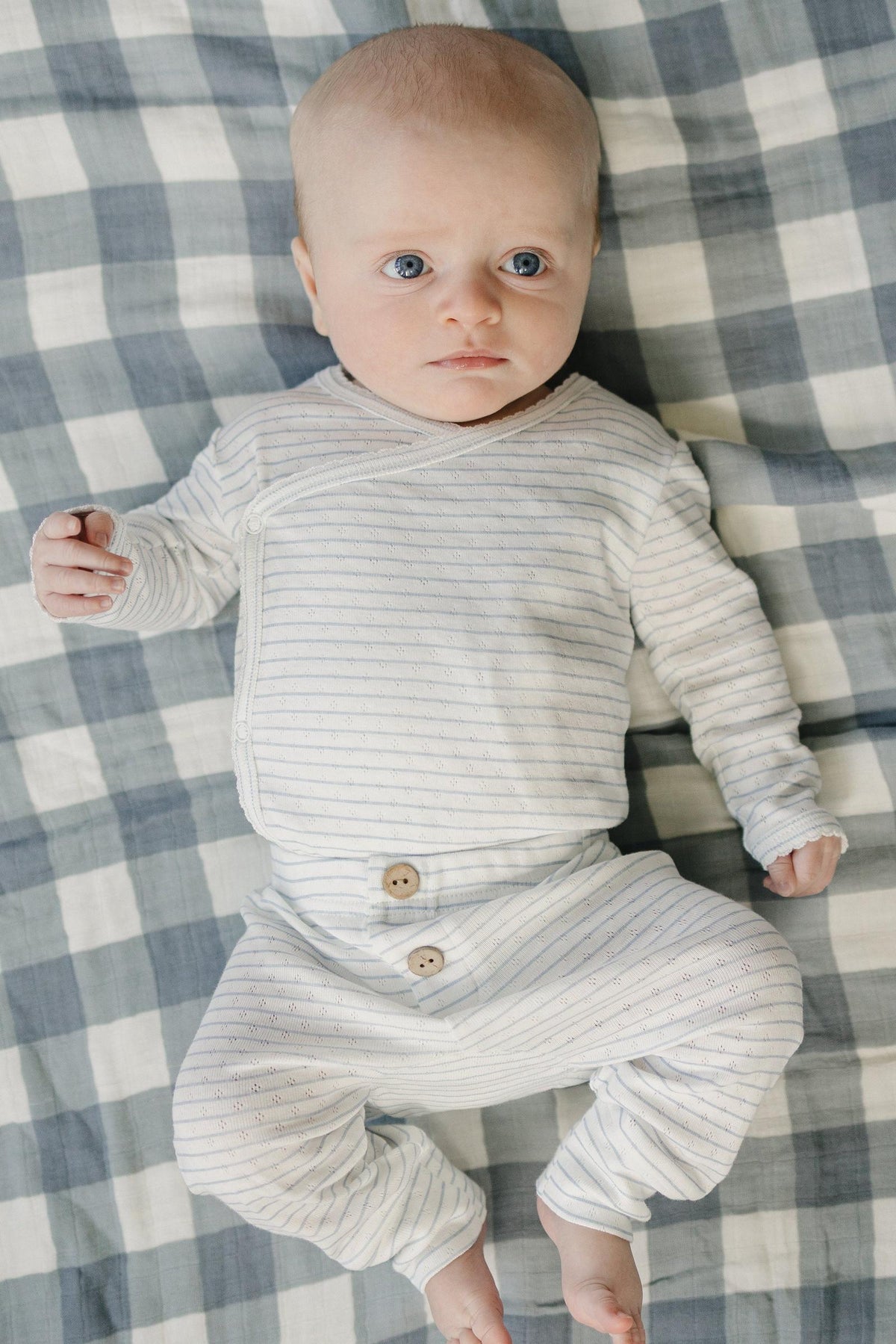 A baby lies on a checkered blanket, wearing a light blue and white striped pointelle layette set with wooden buttons. The outfit is soft and designed for newborn comfort.