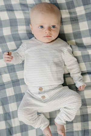 A baby lies on a checkered blanket, wearing a light blue and white striped pointelle layette set with wooden buttons. The outfit is soft and designed for newborn comfort.
