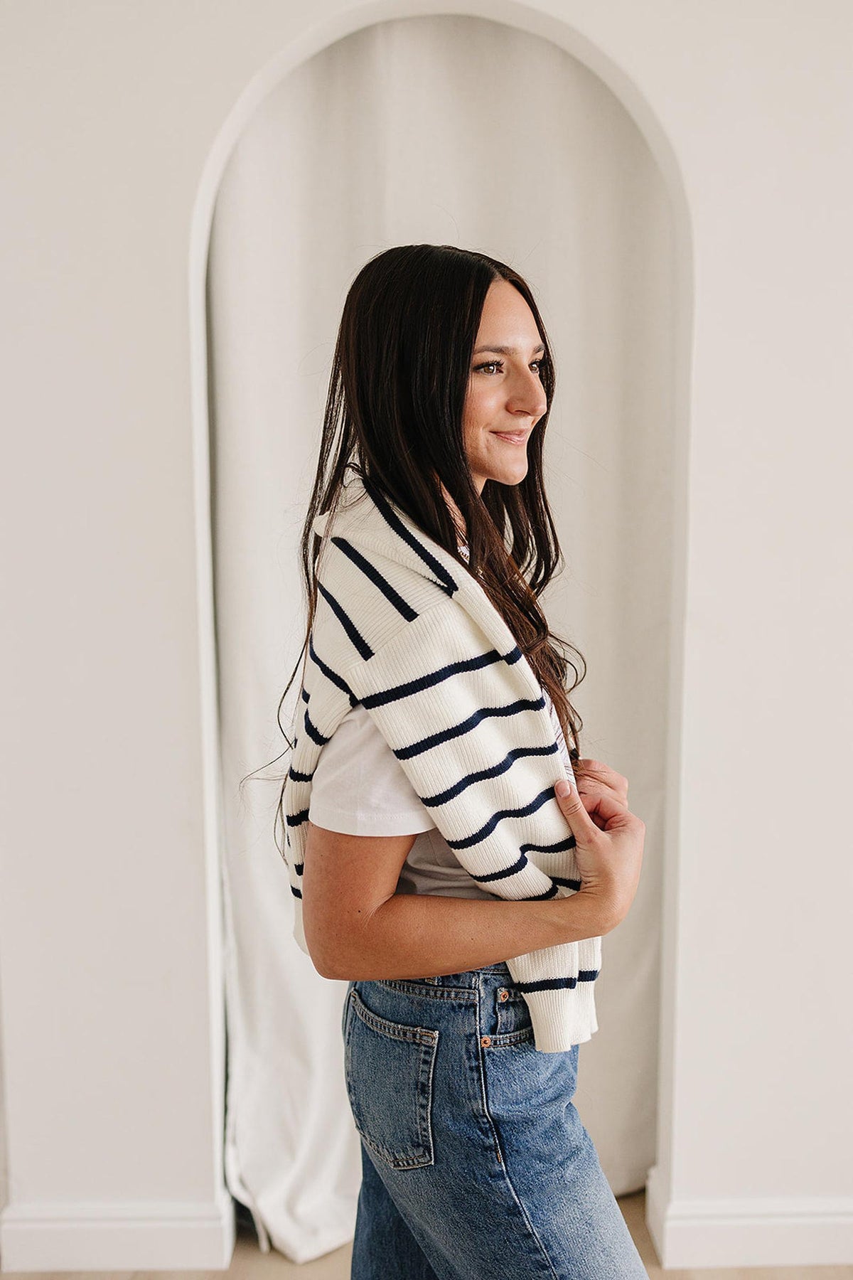 A woman wearing a white shirt and jeans drapes a navy striped sweater over her shoulders in a bright room.