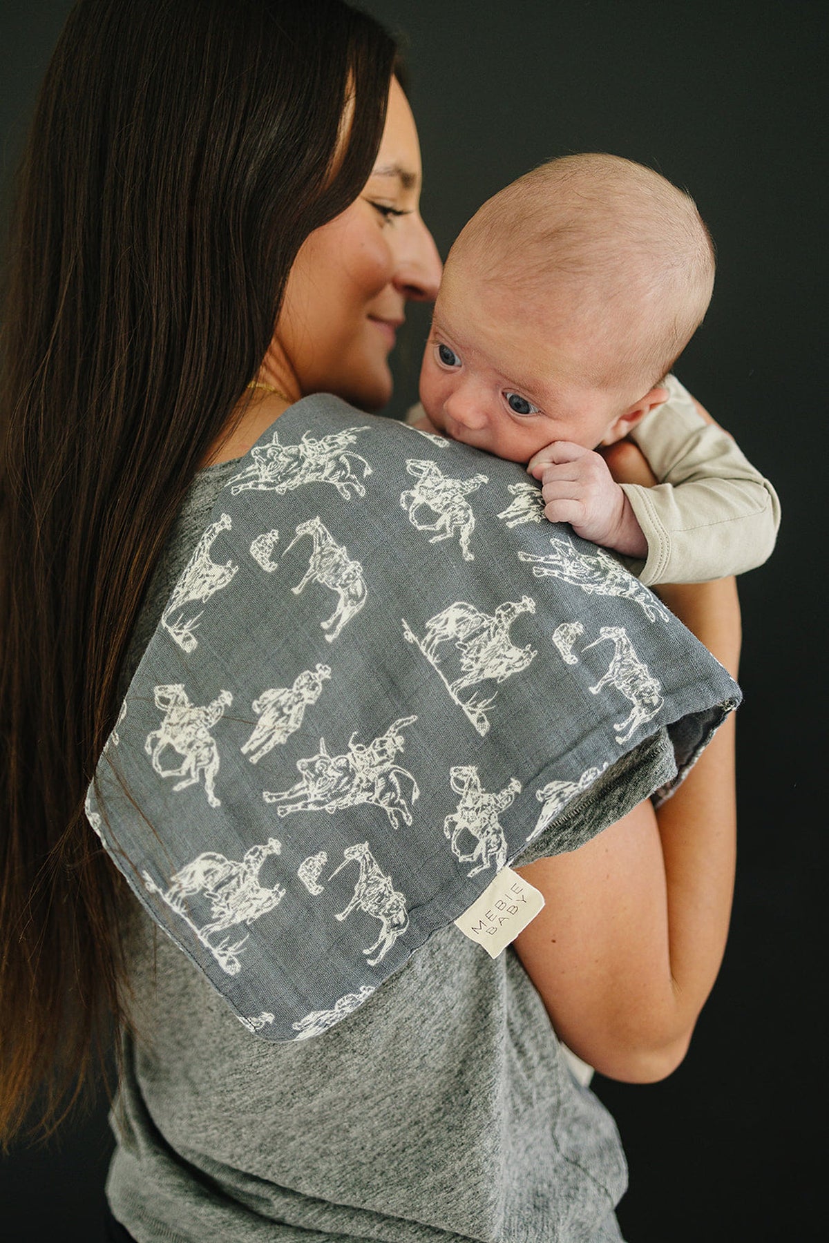 A woman holds a baby while wearing a gray shirt, featuring a Wild West-themed muslin burp cloth with white patterns.
