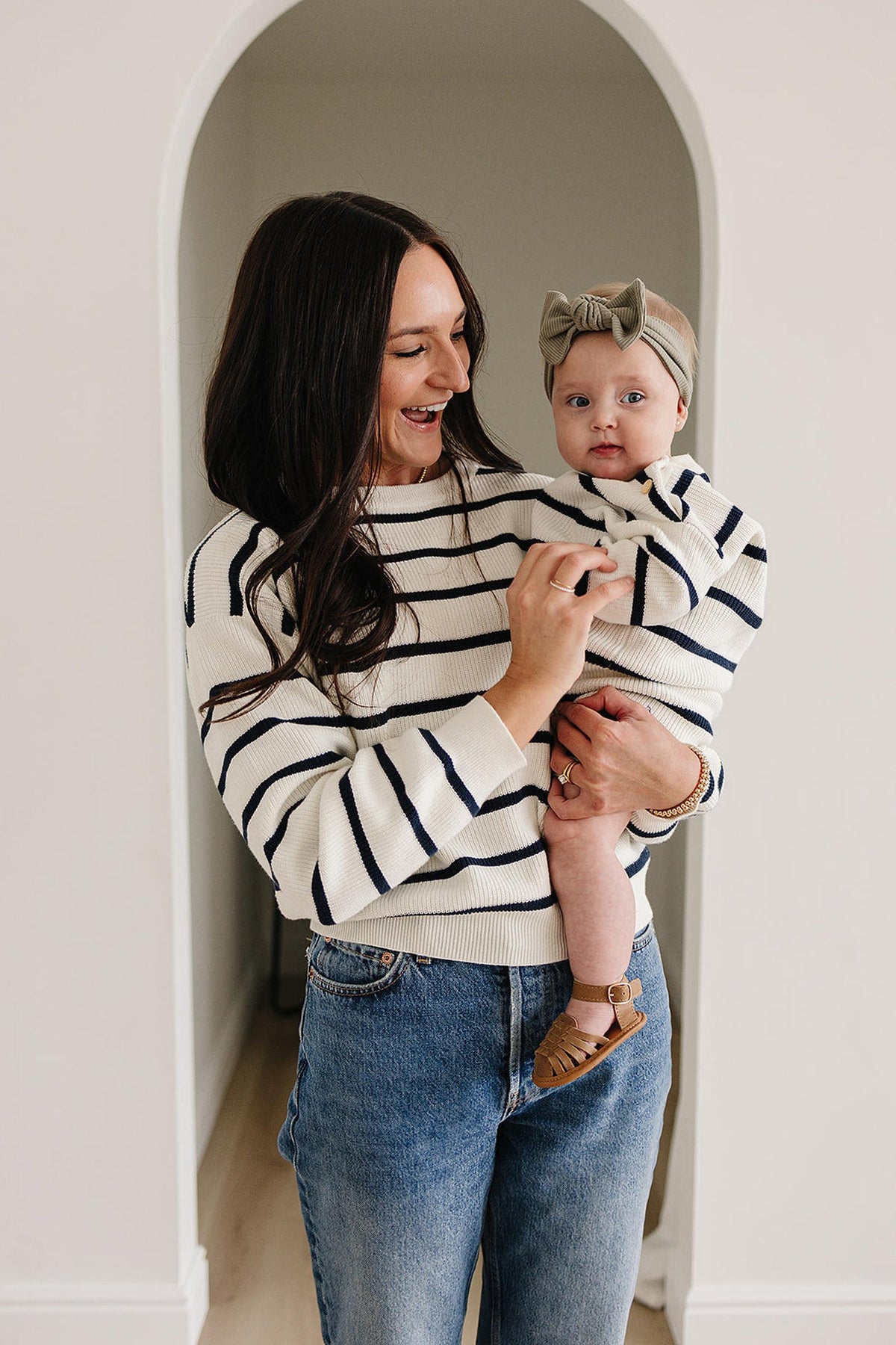 A woman wearing a navy-striped sweater holds a baby dressed similarly. The baby has a bow headband.
