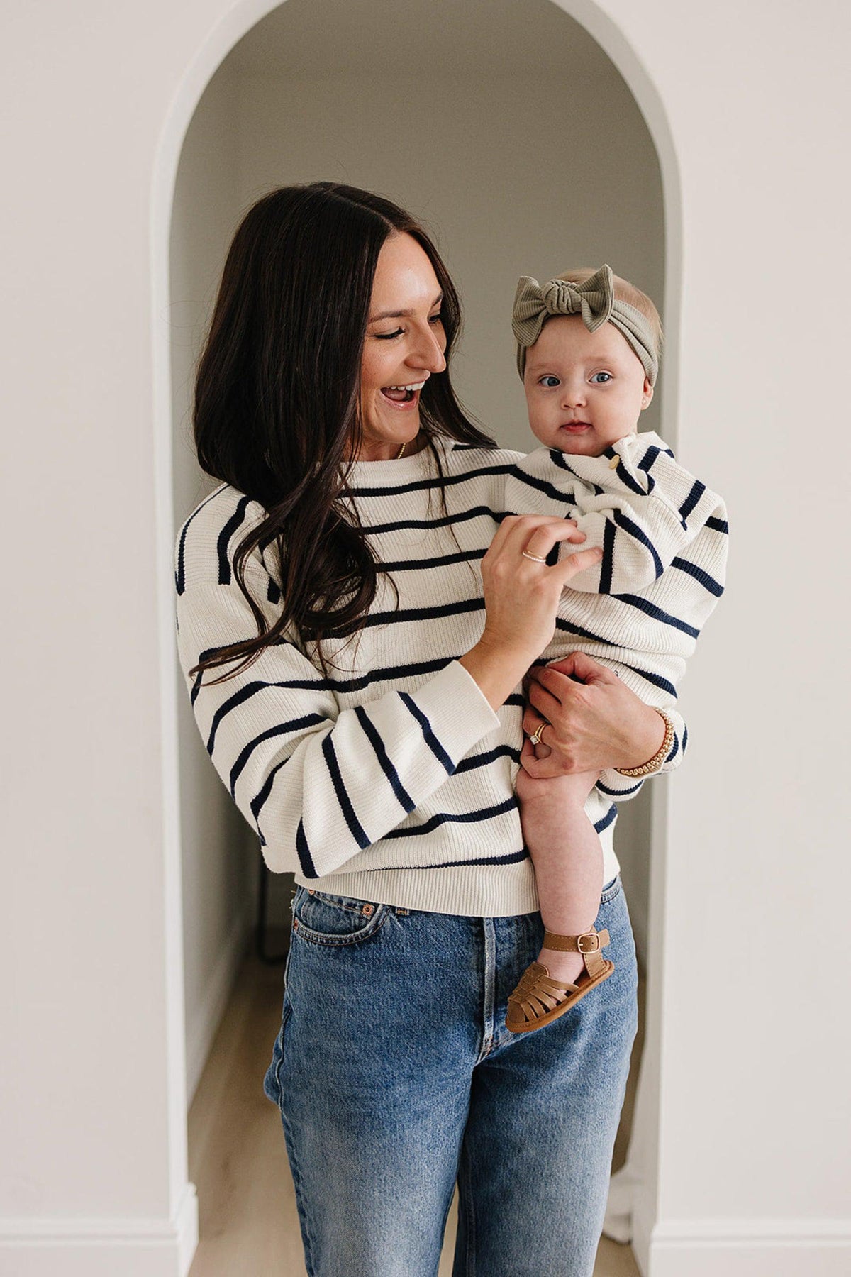 A woman holds a baby wearing matching navy and white striped sweaters in a bright, arched doorway.