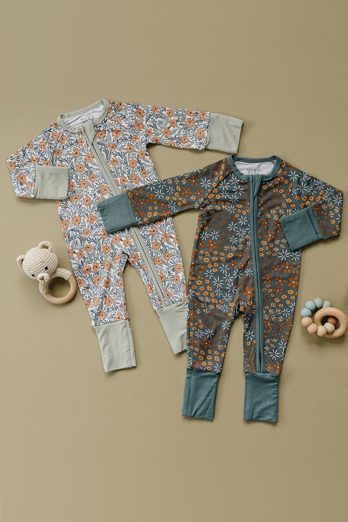 Two floral patterned baby pajamas in soft colors, featuring zippers and foldable mittens, with a plush bear and teething rings.