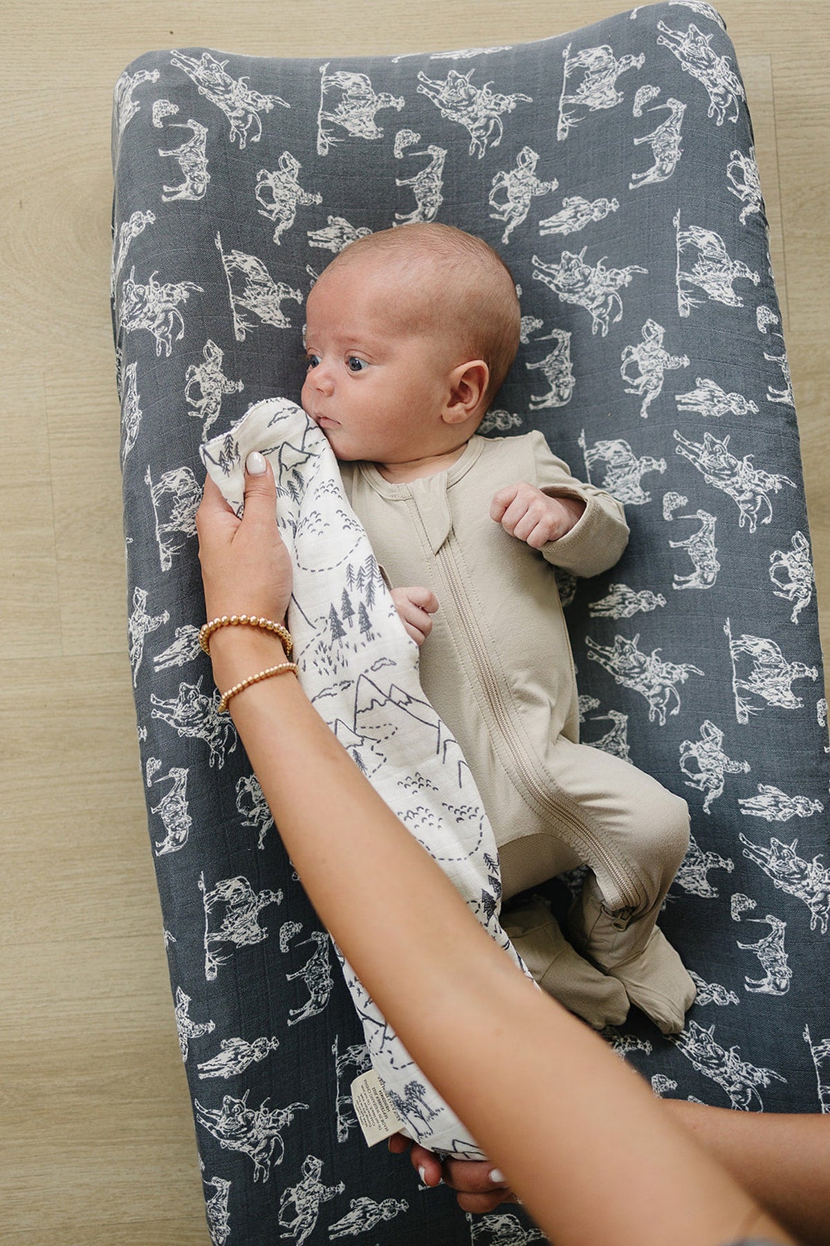 A baby in beige sleepwear lies on a patterned cushion while a hand holds a mountain print muslin cloth.