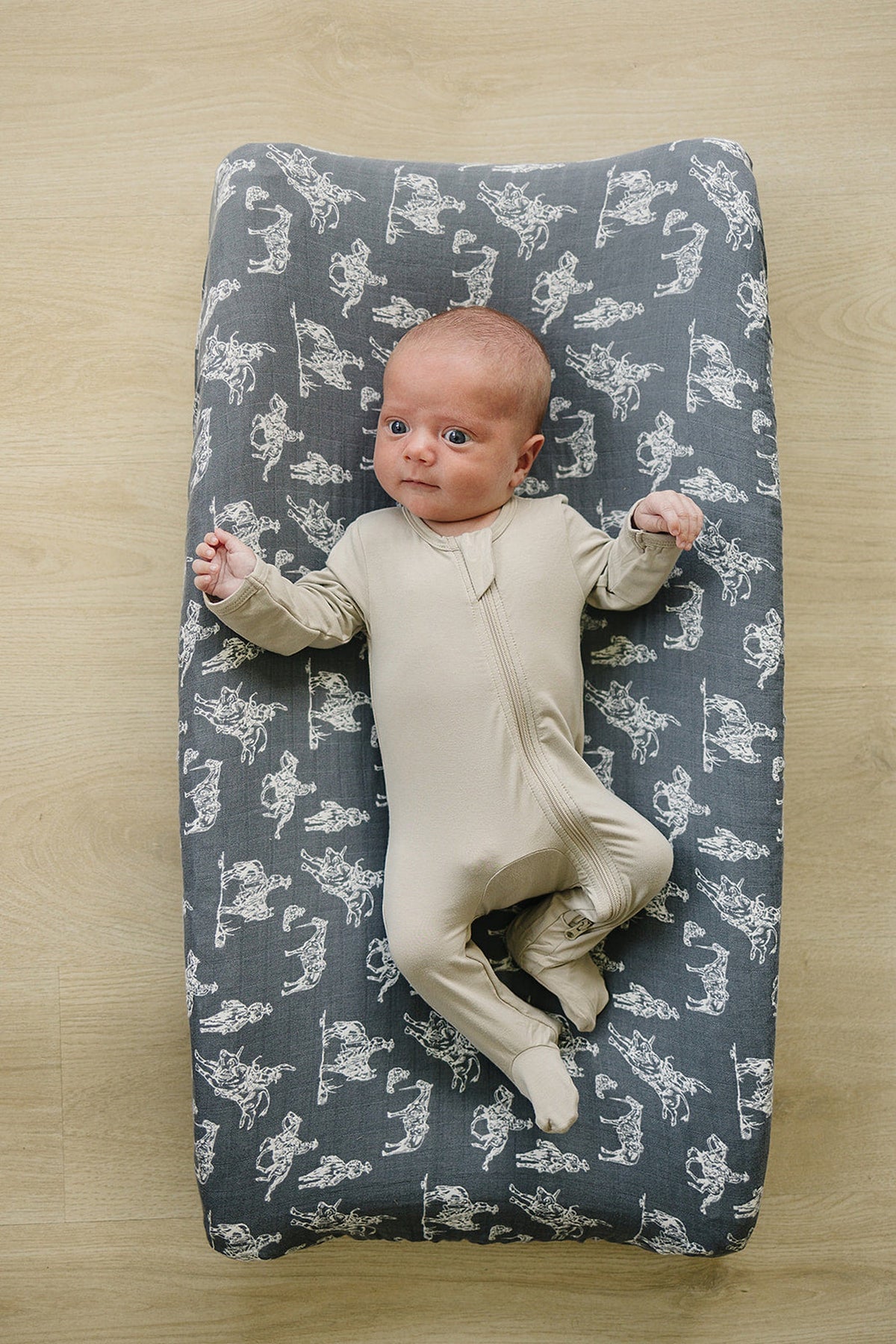 A baby lies on a grey muslin changing pad cover with white Wild West illustrations, wearing a beige onesie.