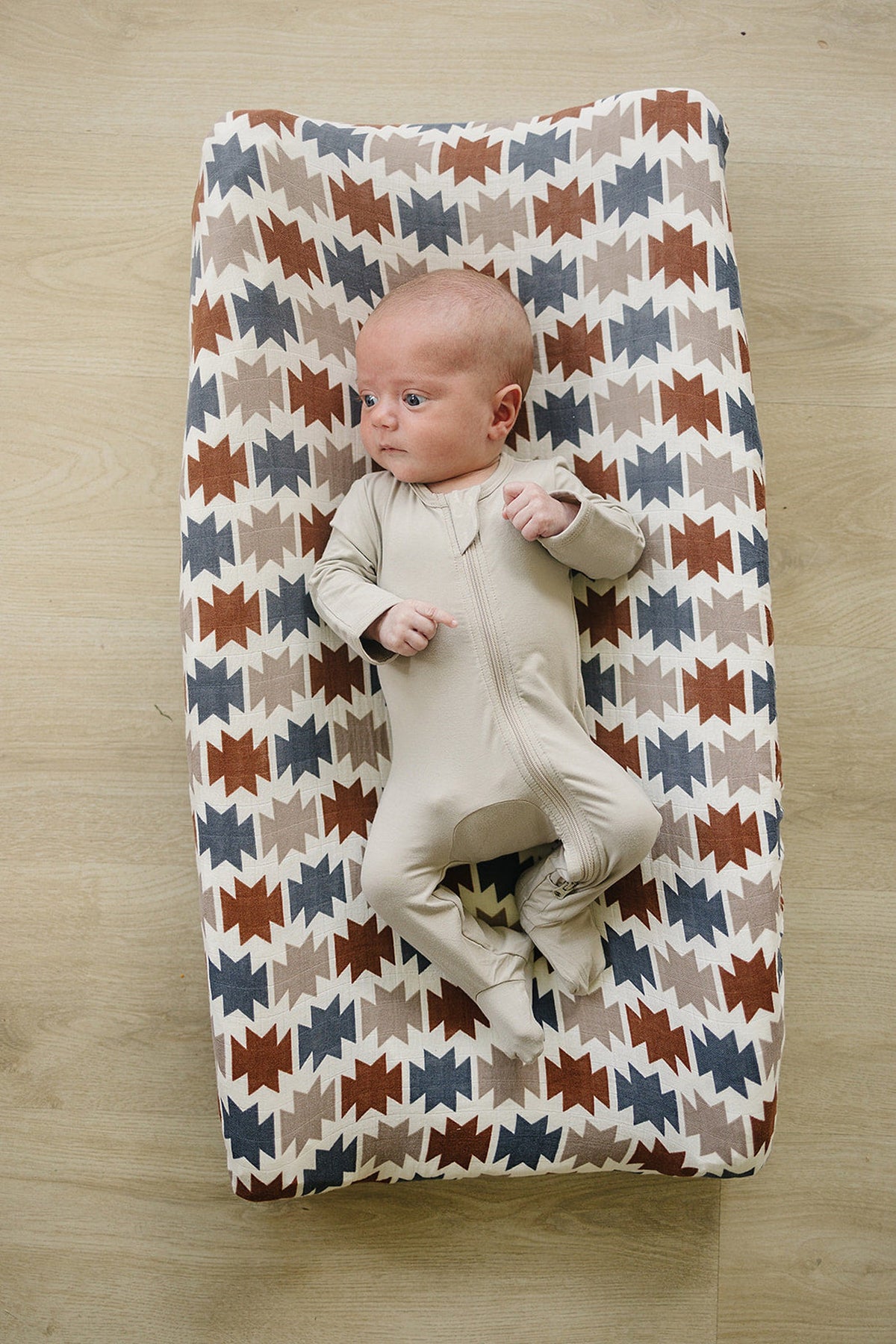 A baby in a beige onesie lies on a changing pad with a Western Aztec pattern in earthy tones.