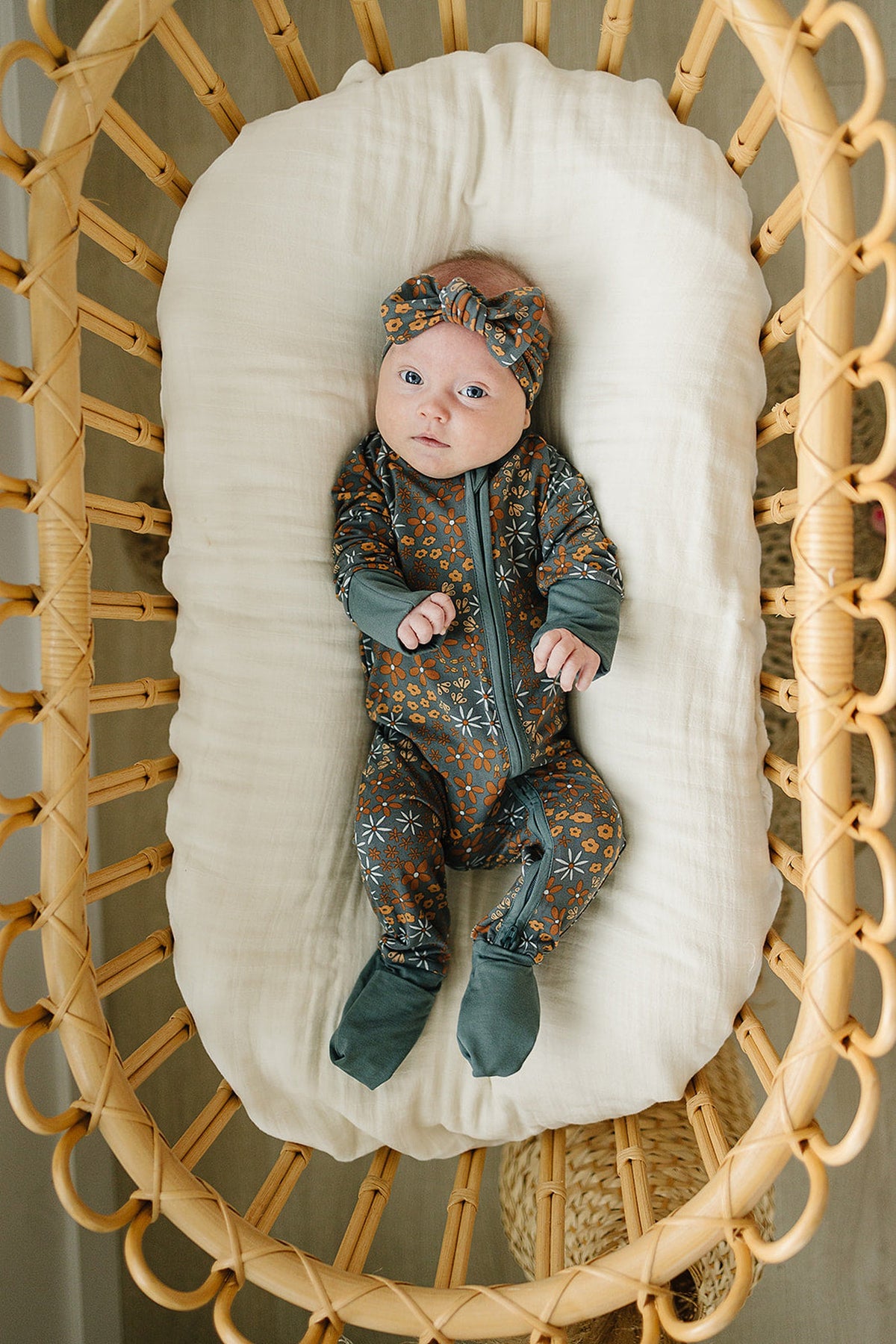 A baby wearing a floral patterned outfit and head wrap lies in a wicker bassinet on a white cushion.