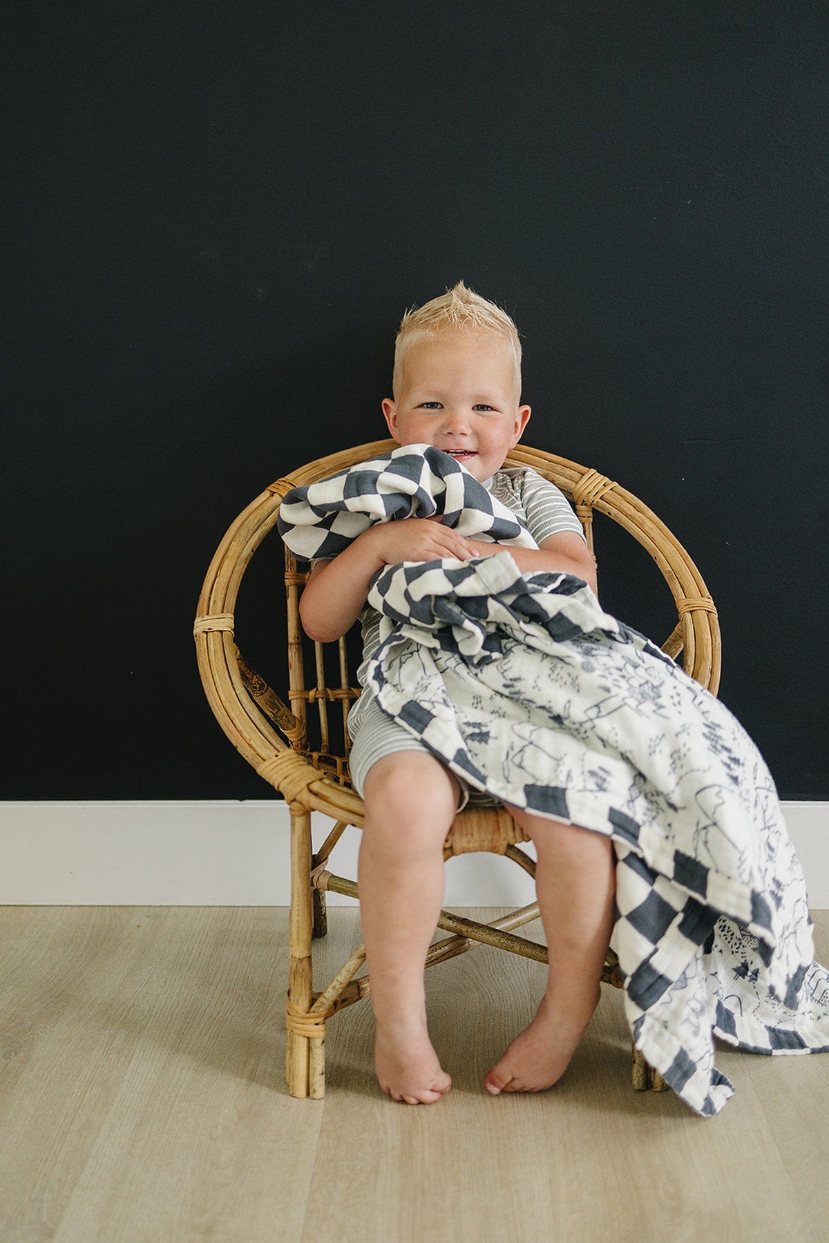 A smiling child sits on a rattan chair, holding a charcoal checkered muslin quilt against a dark wall backdrop.