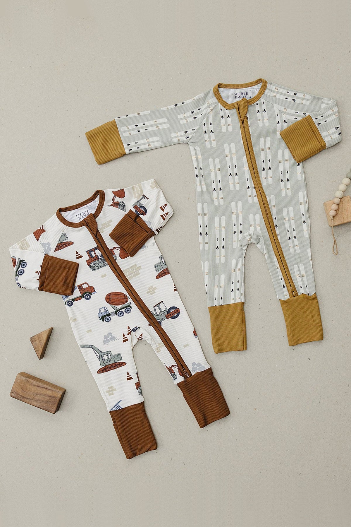 Two Mebie Baby ribbed zipper pajamas, one with construction trucks and the other with geometric patterns, on a beige background.