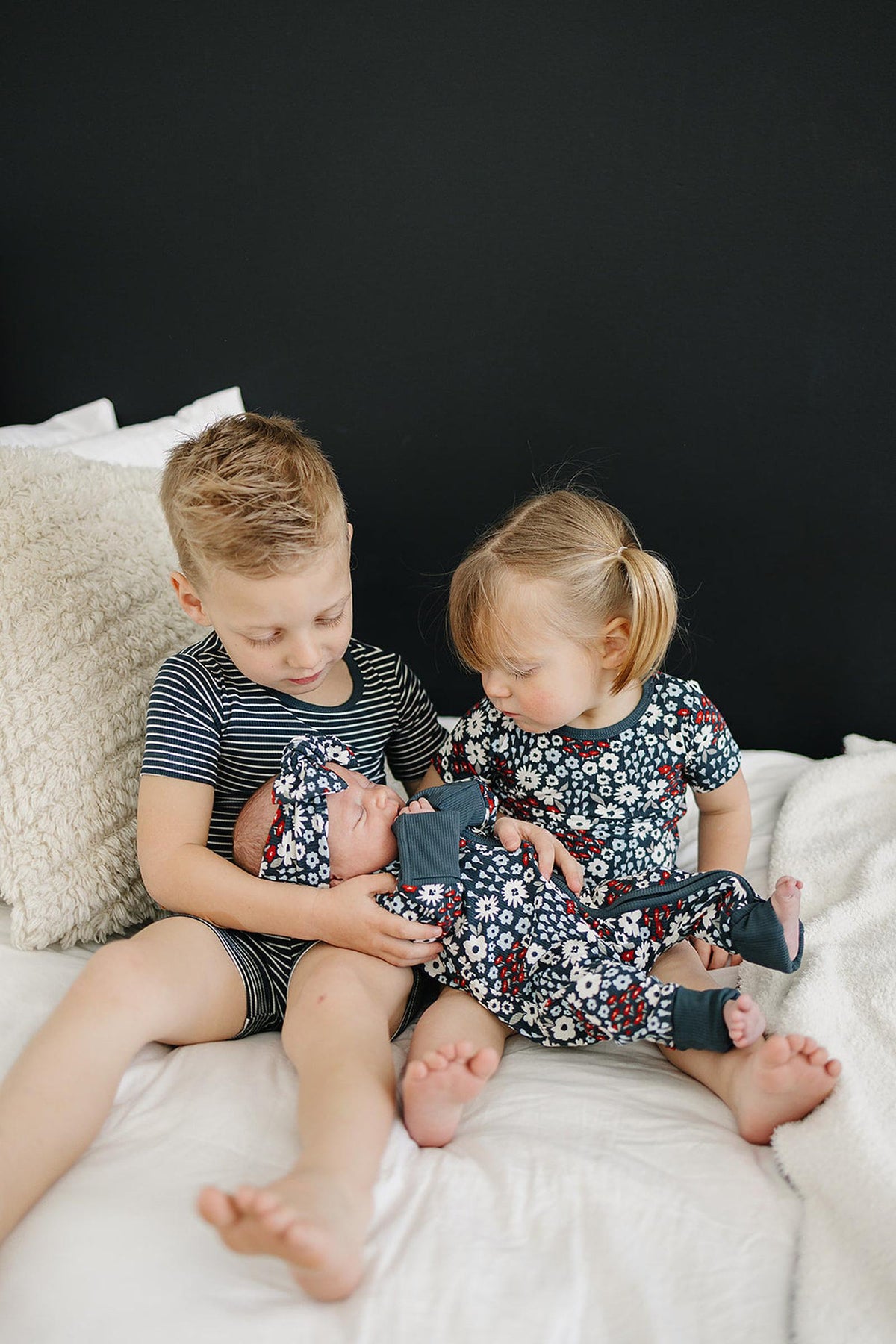 Two young children sitting on a bed, holding a baby in floral pajamas and headband against a dark background.