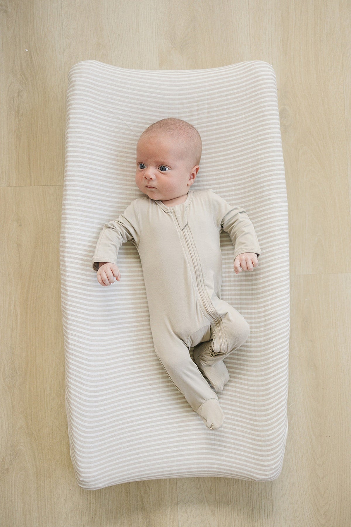A baby in a beige onesie lies on a taupe striped changing pad cover on a wooden floor.