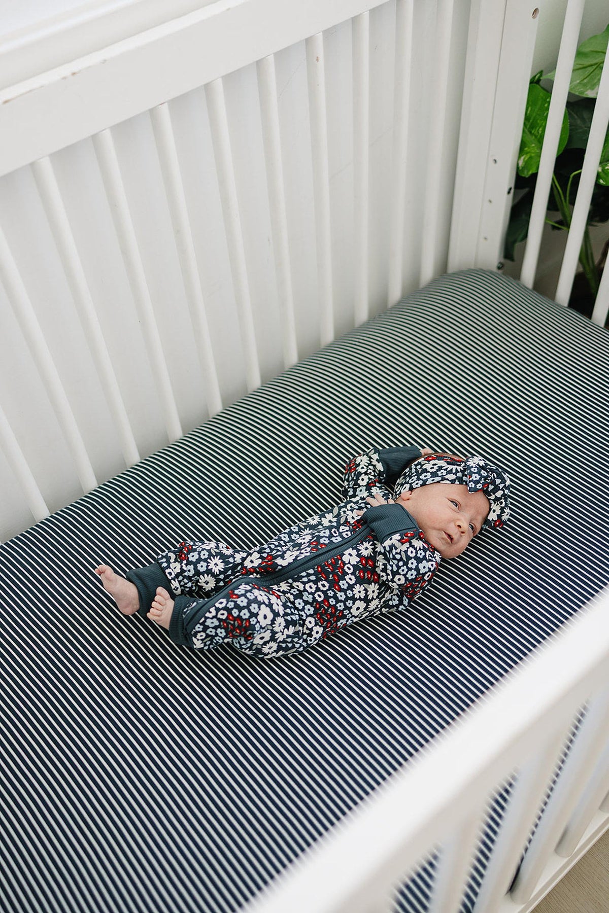 A baby in a floral onesie lies on a navy striped crib sheet in a white crib.