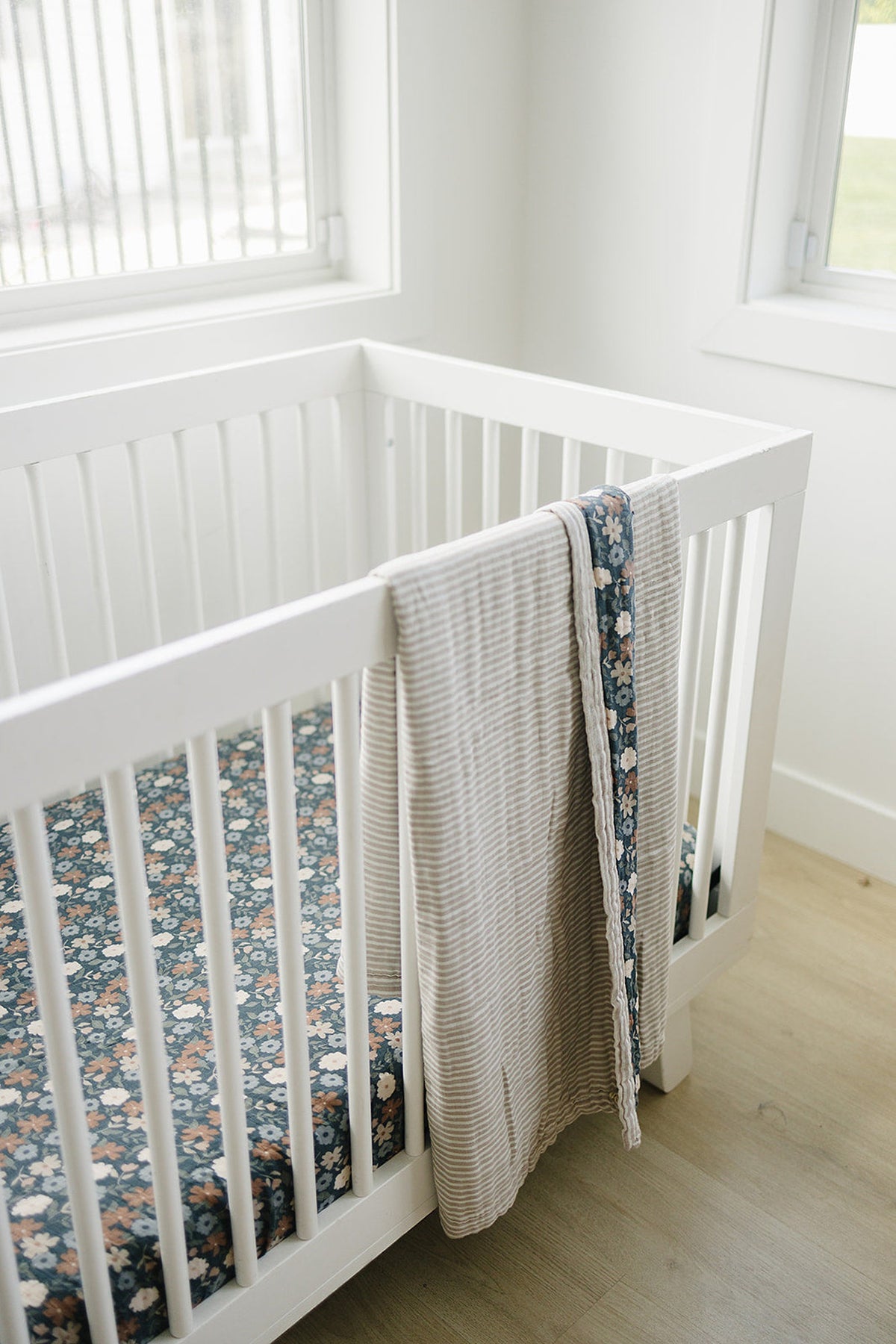 A white crib with a floral muslin crib sheet featuring dark blue and pastel flowers, paired with a striped beige blanket draped over the side, set in a bright, airy.