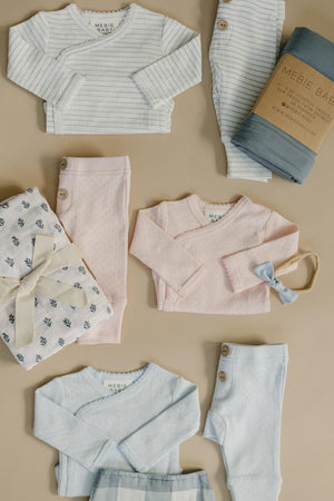 A collection of Mebie Baby layette sets features soft, breathable cotton in pastel colors, including pink, blue, and white stripes. Each outfit includes gentle stretch for newborn comfort and style.