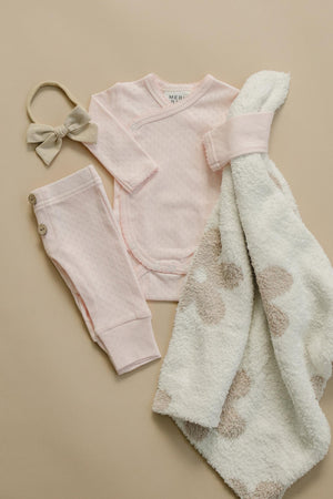 A soft pink pointelle layette set by Mebie, featuring a long-sleeve wrap shirt, matching pants, a bow headband, and a cozy white towel with light brown floral patterns.
