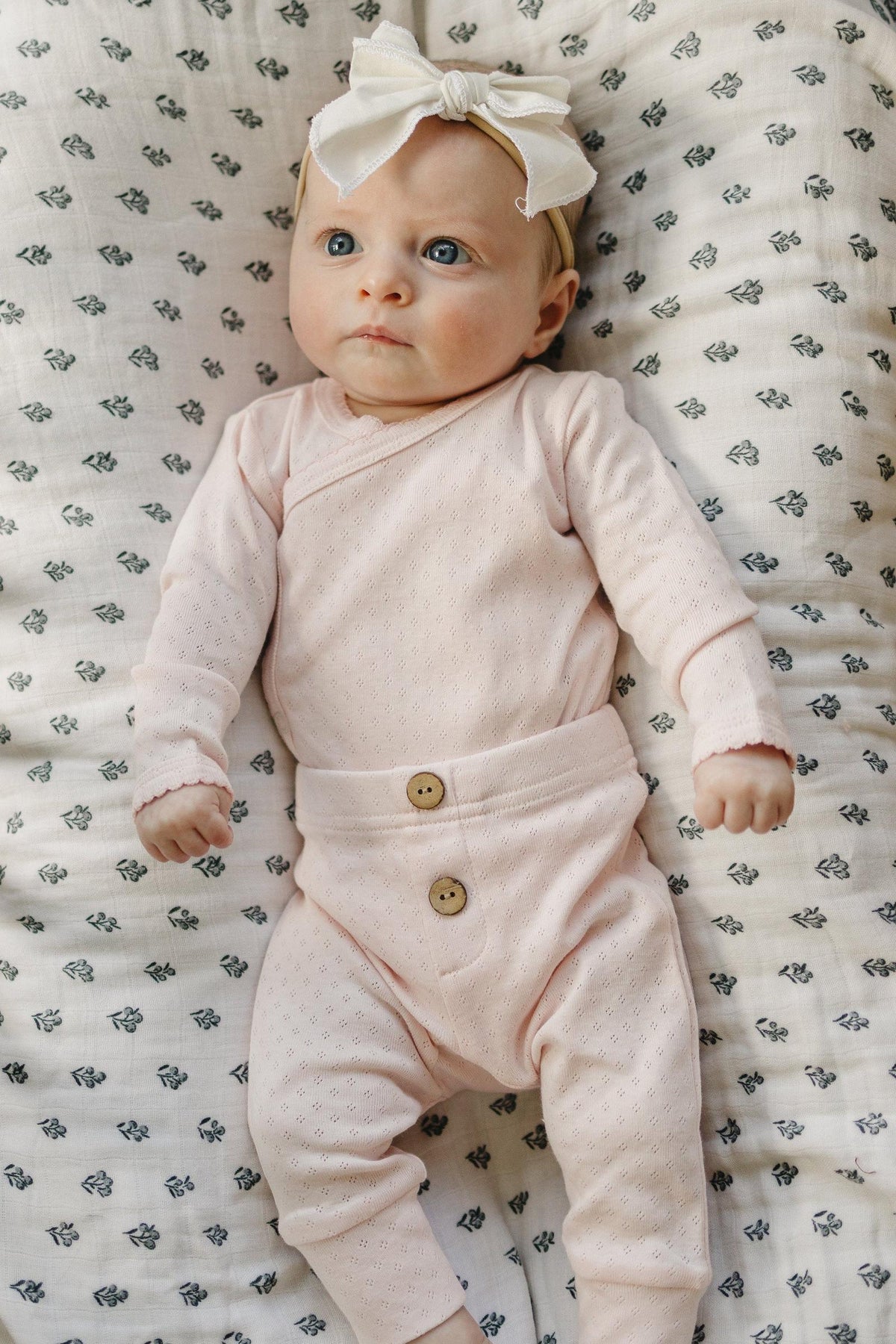 A baby girl lies on a floral-patterned blanket, wearing a soft pink pointelle layette set with a stylish bow headband. The outfit features delicate buttons and comfortable, breathable cotton fabric.