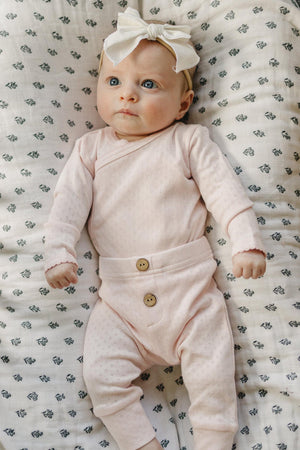 A baby girl lies on a floral-patterned blanket, wearing a soft pink pointelle layette set with a stylish bow headband. The outfit features delicate buttons and comfortable, breathable cotton fabric.