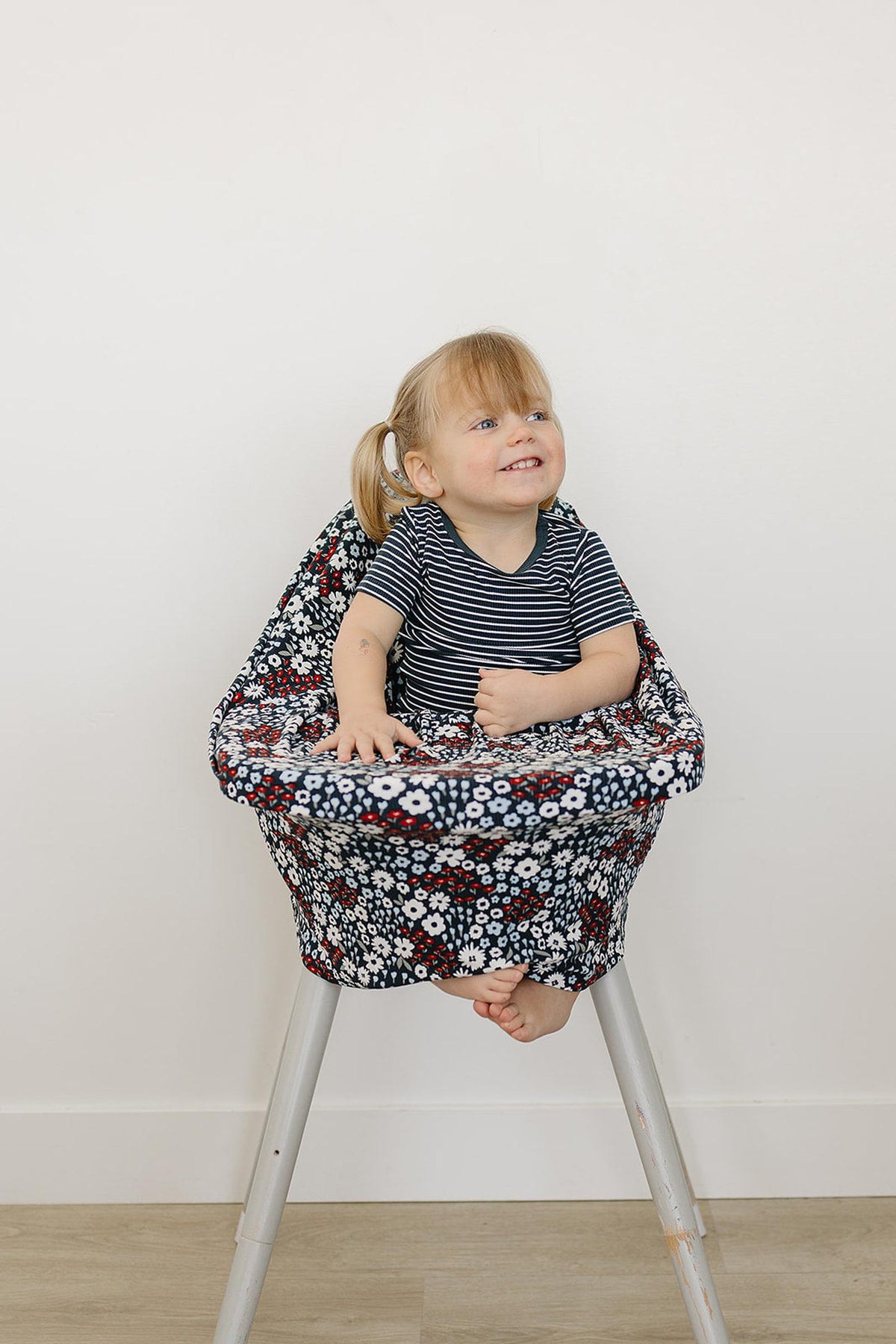 A smiling child sits in a high chair covered with a floral-patterned fabric, wearing a striped shirt.