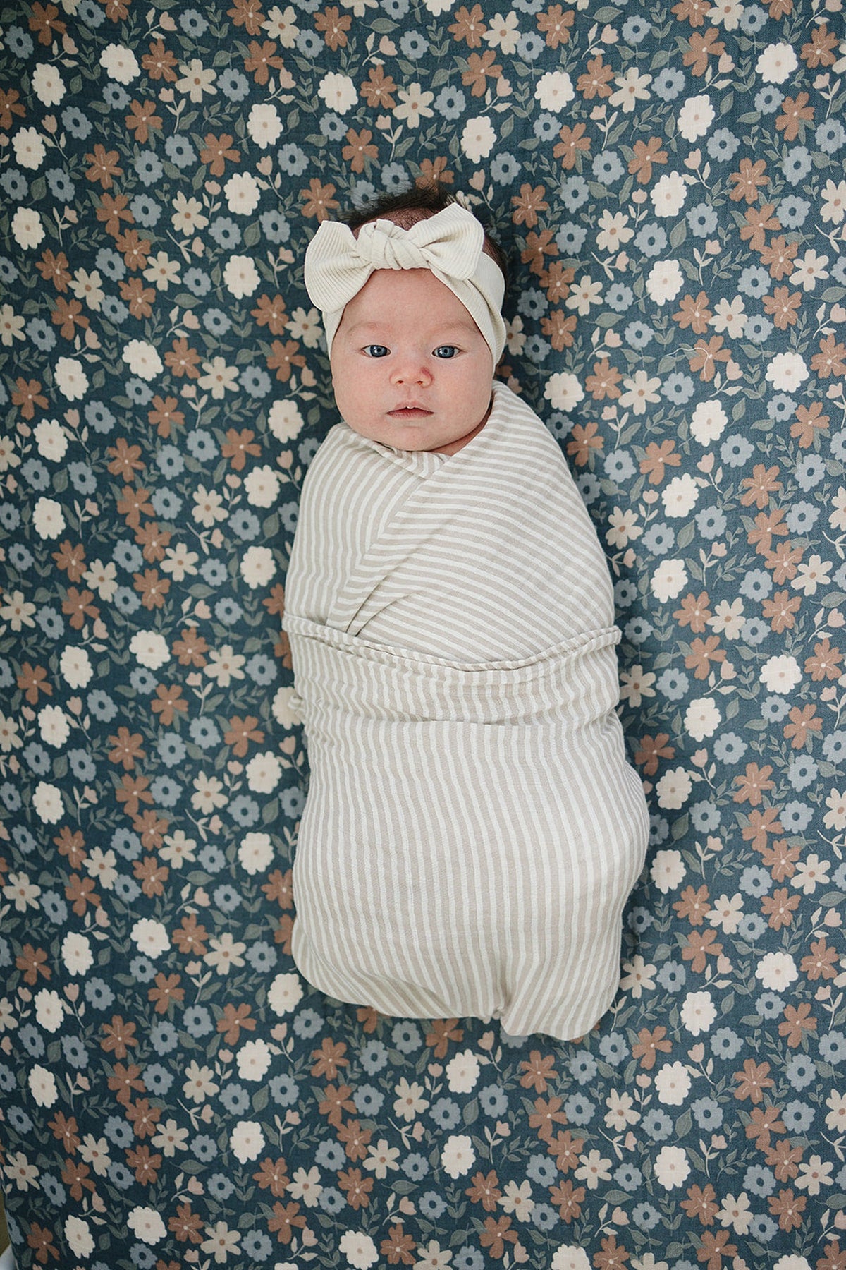 A swaddled baby with a striped fabric is centered on a floral-patterned crib sheet featuring dark blue and multicolored flowers. The baby wears a bow headband, adding a charming touch.