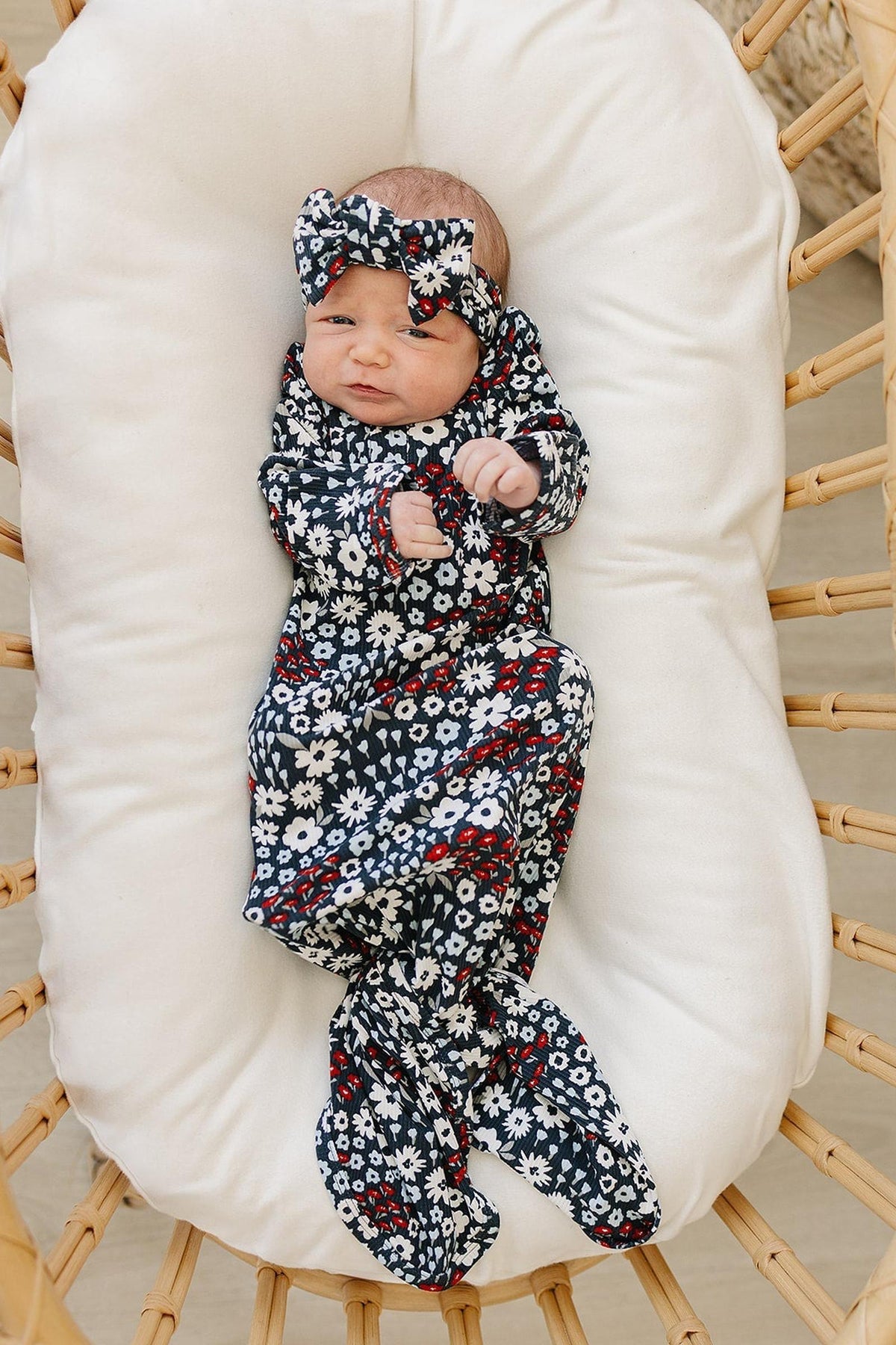 A baby wearing a floral-patterned head wrap and matching outfit lies on a cushioned wicker chair.