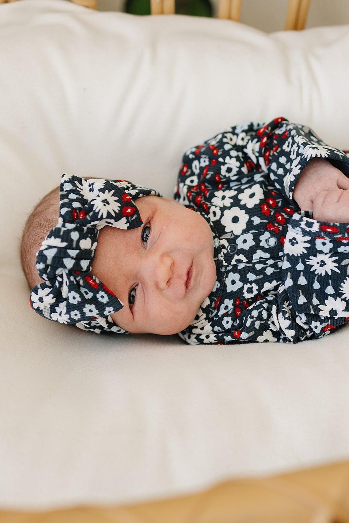 A baby wearing a floral-patterned head wrap and outfit lies on a cream-colored cushion.