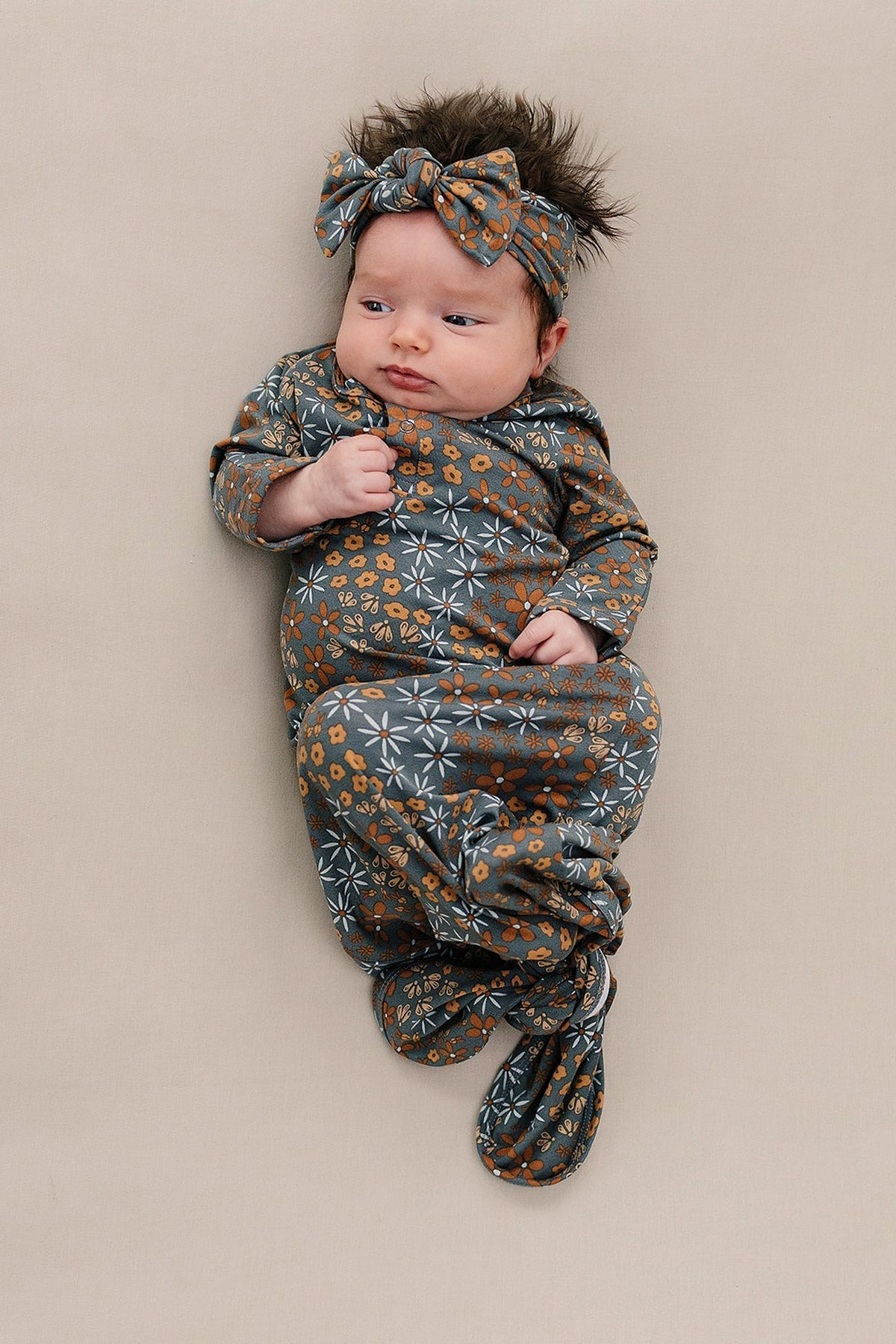 A baby wearing a floral patterned sleeper and matching headband lies on a beige surface, looking to the side.