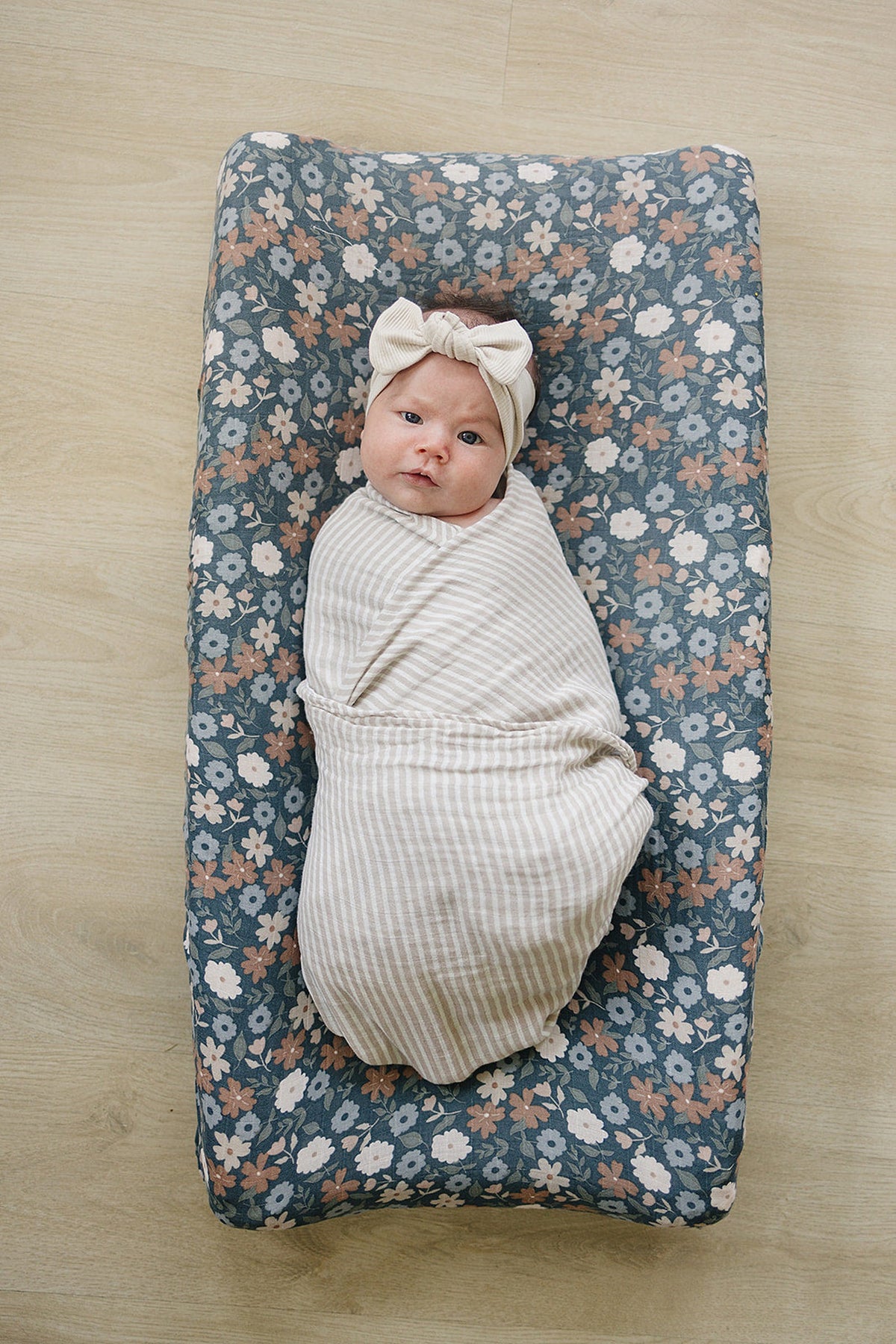 A swaddled baby lies on a floral muslin changing pad cover, featuring a blue background with pink and white flowers. The baby wears a light-colored headband, adding a charming touch.