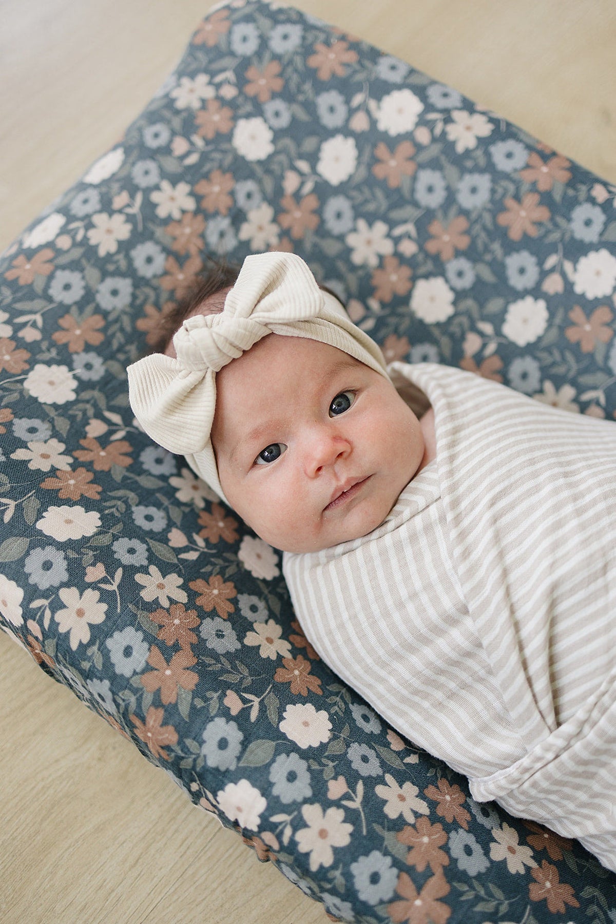 A baby wrapped in a striped swaddle lies on a midnight floral muslin changing pad cover with soft, breathable fabric.