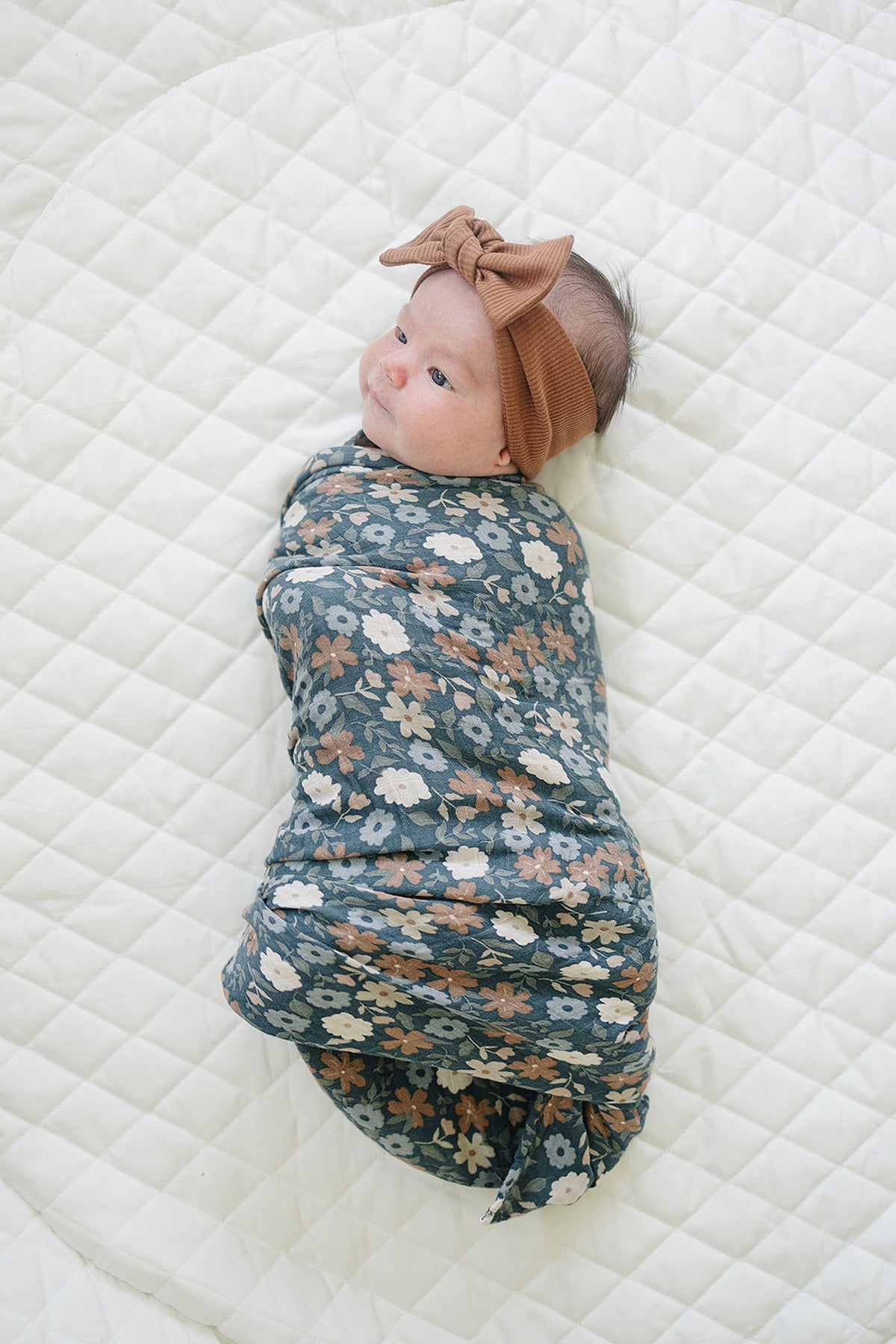 A baby is wrapped in a navy floral muslin swaddle blanket, lying on a white quilted surface.
