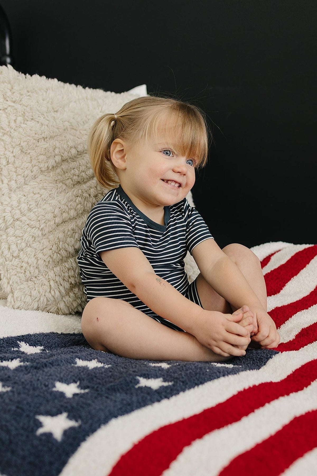 A toddler in a navy and white striped outfit sits on a star-spangled blanket, smiling.