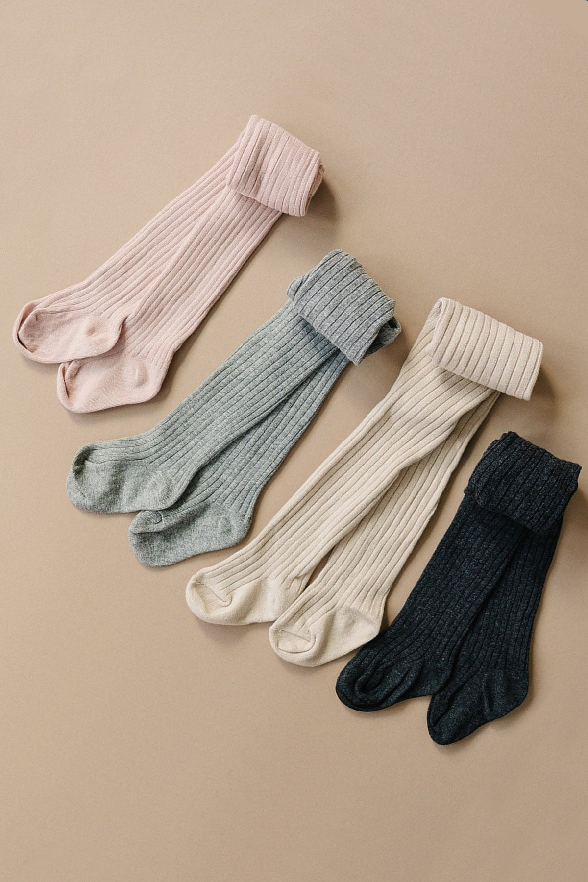 Four pairs of ribbed baby tights in pink, blue, cream, and black are neatly arranged on a beige background.