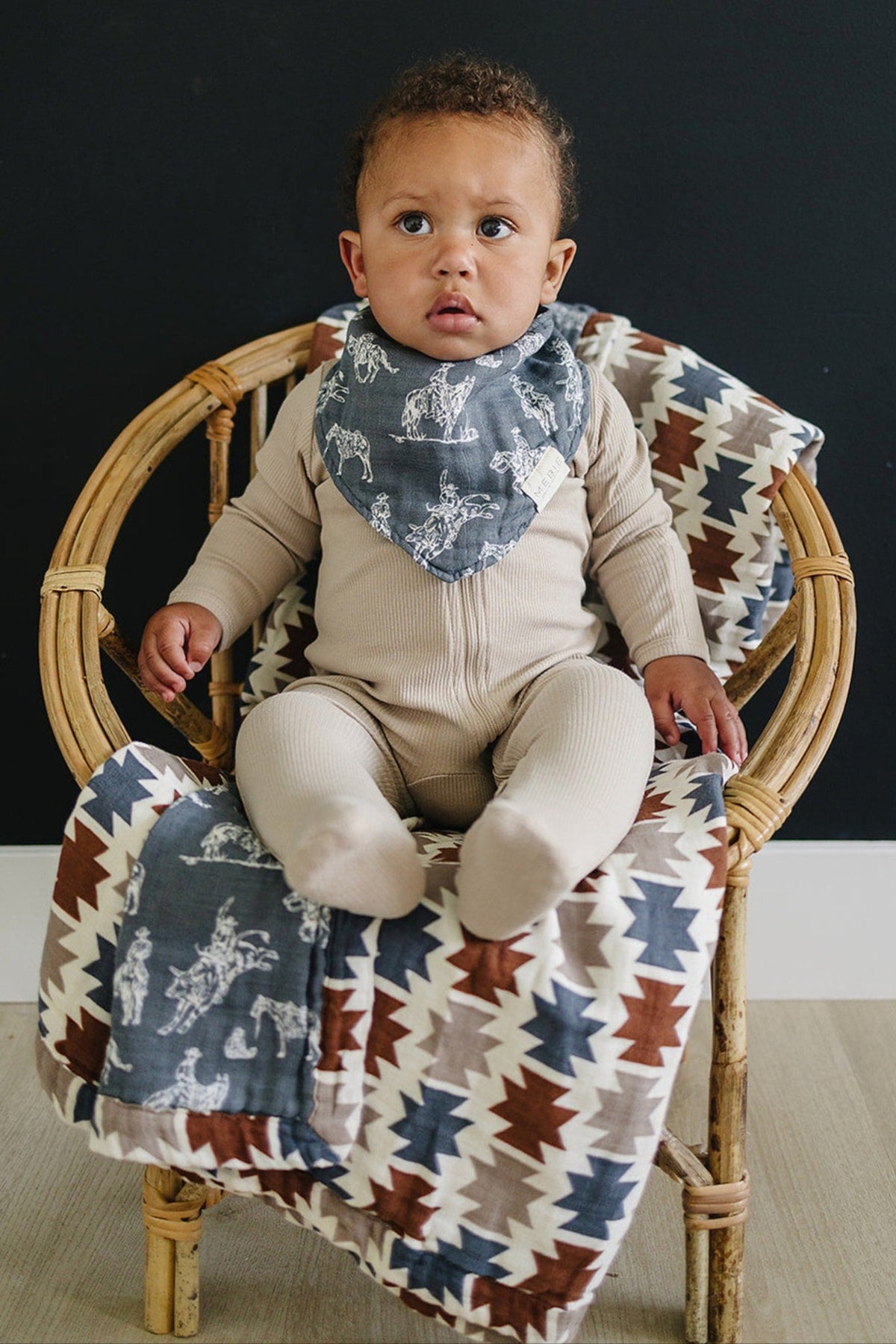 A baby wearing a Mebie Baby Wild West Muslin Bib sits on a patterned blanket in a wicker chair.