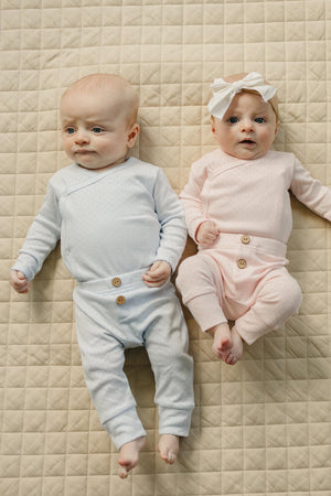 Two infants lie on a quilted beige surface, wearing cozy, pastel-colored layette sets. One baby is in a blue pointelle outfit, while the other wears a soft pink ensemble with.