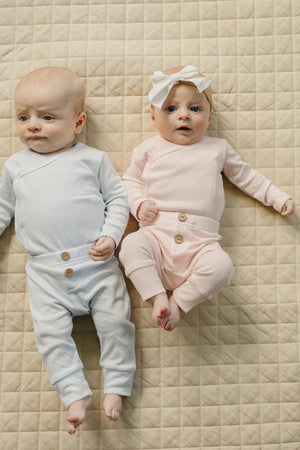 Two infants lie side by side on a beige quilt. One wears a light blue pointelle outfit.