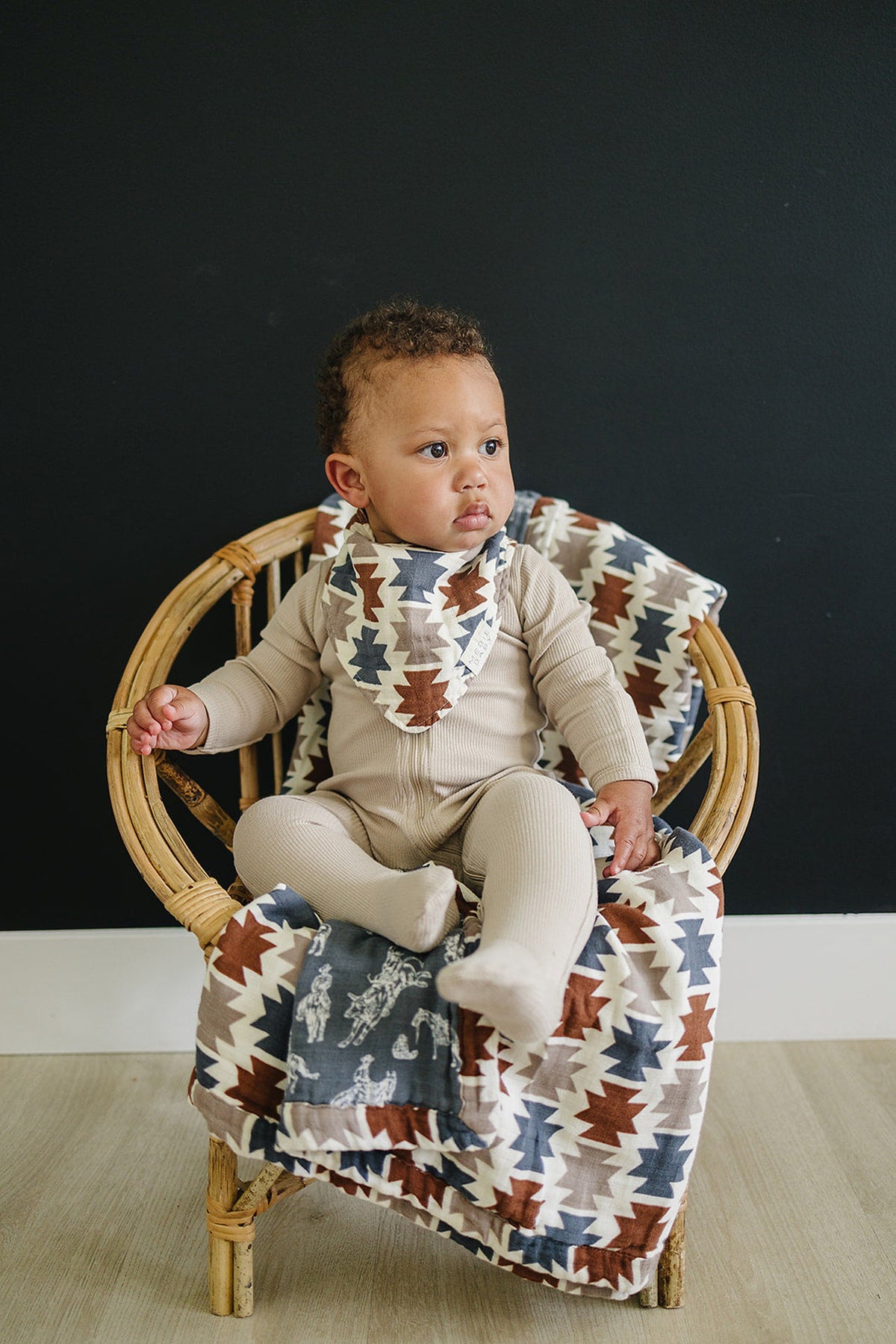 A baby wearing a Western Aztec patterned bib sits on a wooden chair with a matching blanket.