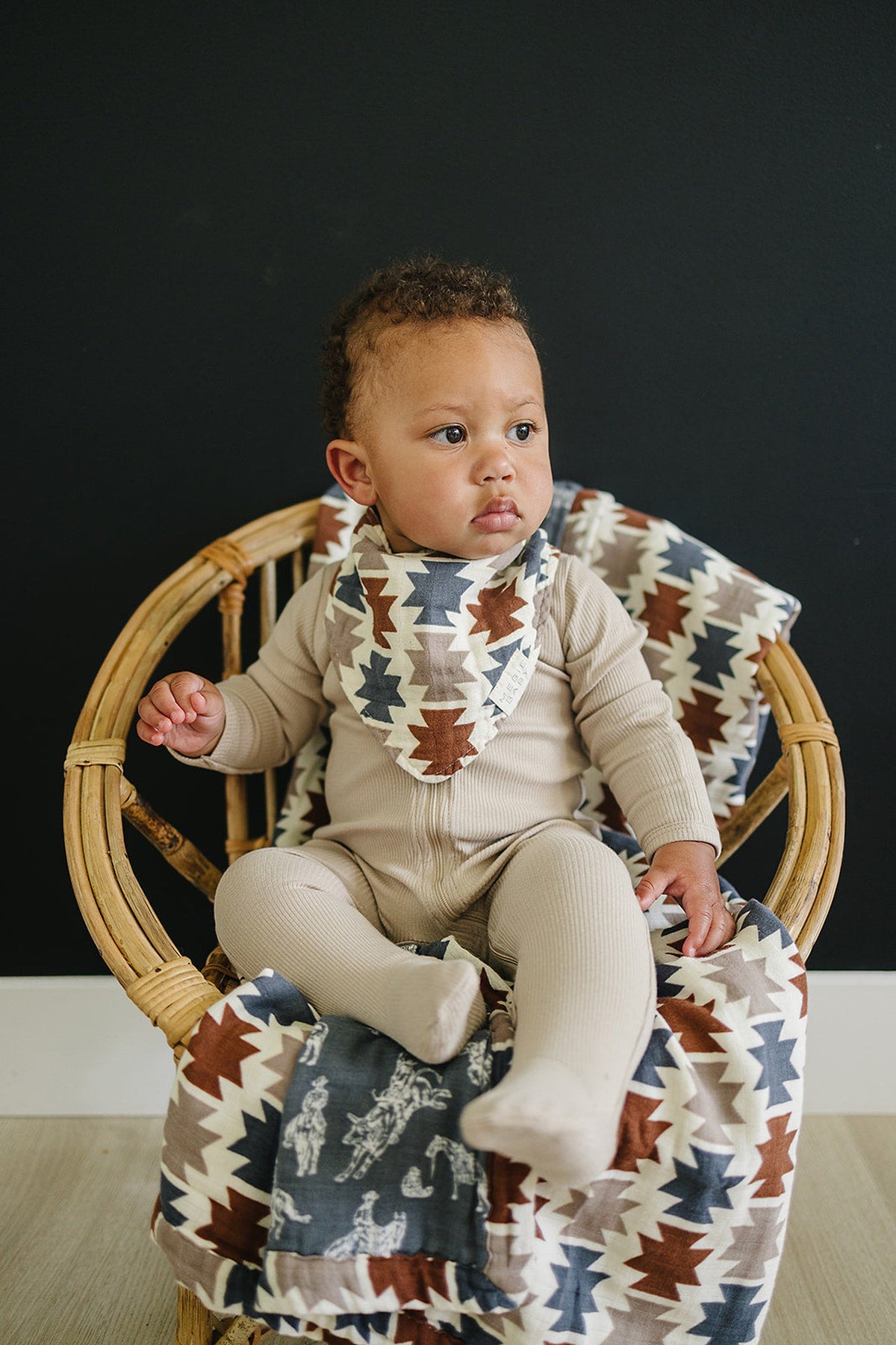A baby in a beige onesie sits on a wooden chair with an Aztec-patterned quilt and bib.