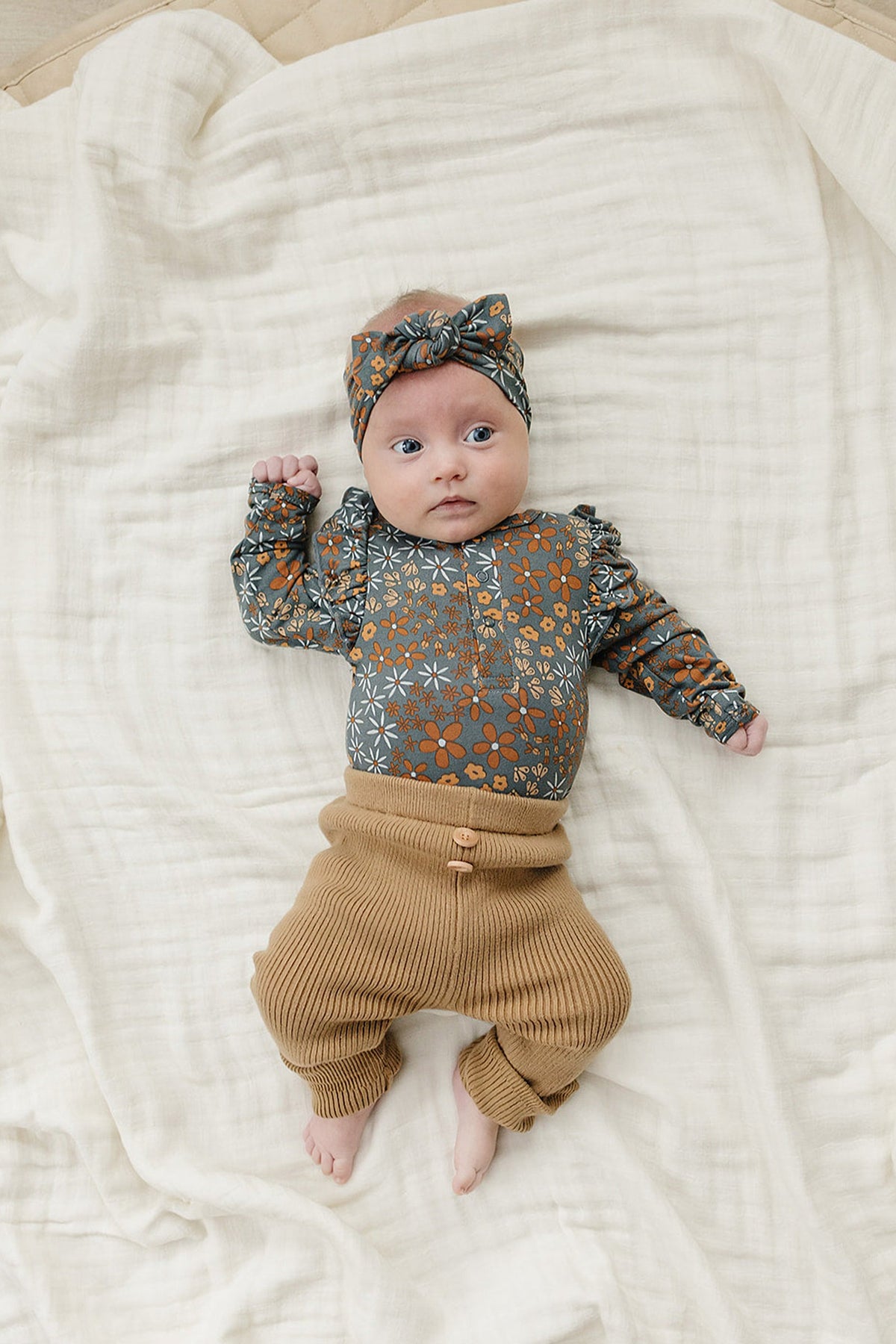 A baby wearing a floral-patterned outfit and head wrap lies on a soft, cream-colored blanket.
