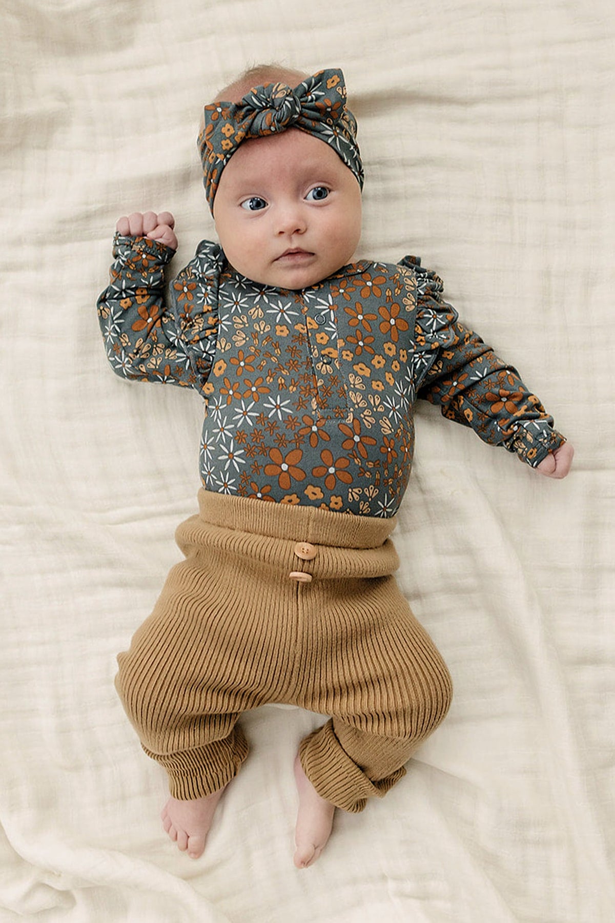 Baby wearing a floral ruffle sleeve bodysuit and headband, paired with ribbed brown pants, lying on a blanket.