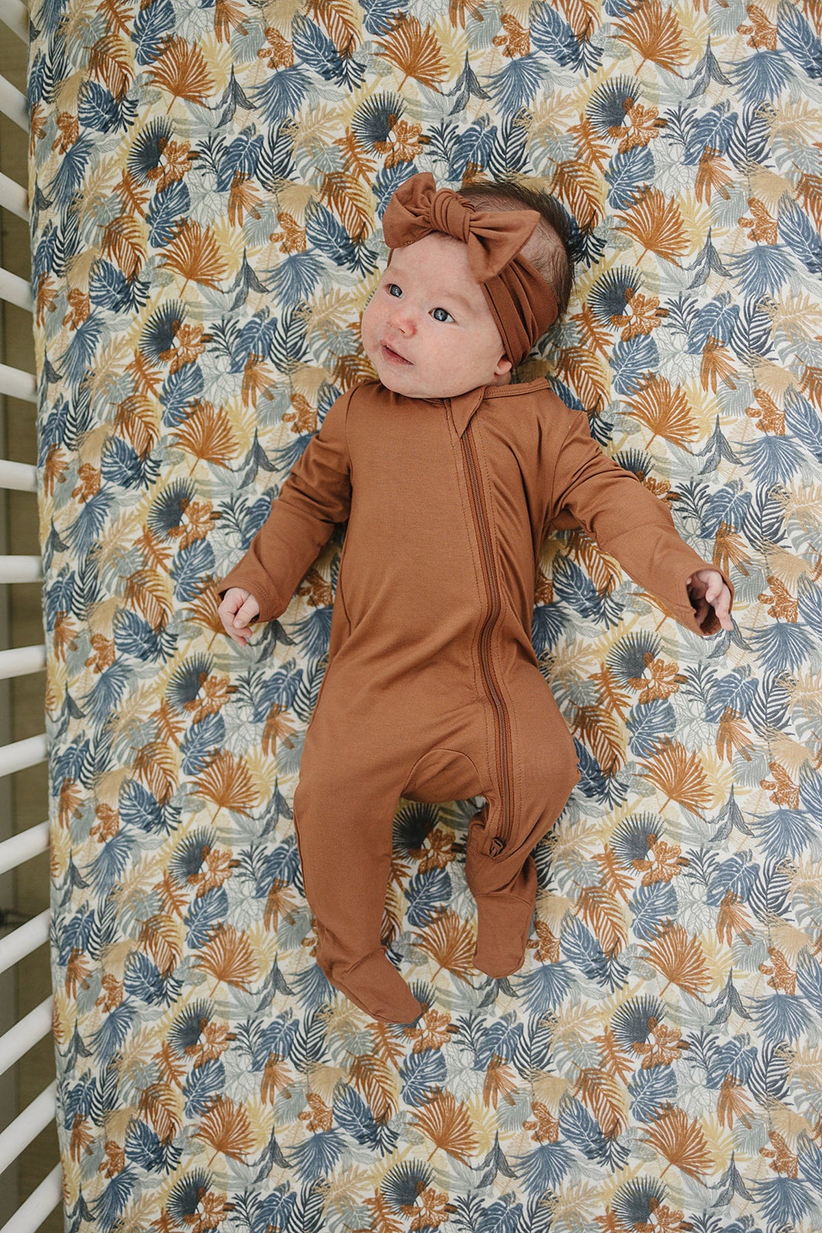 A baby in a brown outfit and headband lies on a tropical leaf-patterned crib sheet.