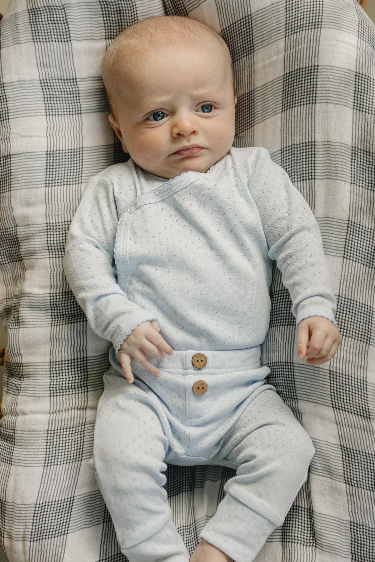 A newborn boy lies on a checkered blanket, wearing a light blue pointelle layette set with two wooden buttons. The outfit features a soft, breathable cotton material, ensuring comfort.