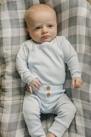 A newborn boy lies on a checkered blanket, wearing a light blue pointelle layette set with two wooden buttons. The outfit features a soft, breathable cotton material, ensuring comfort.
