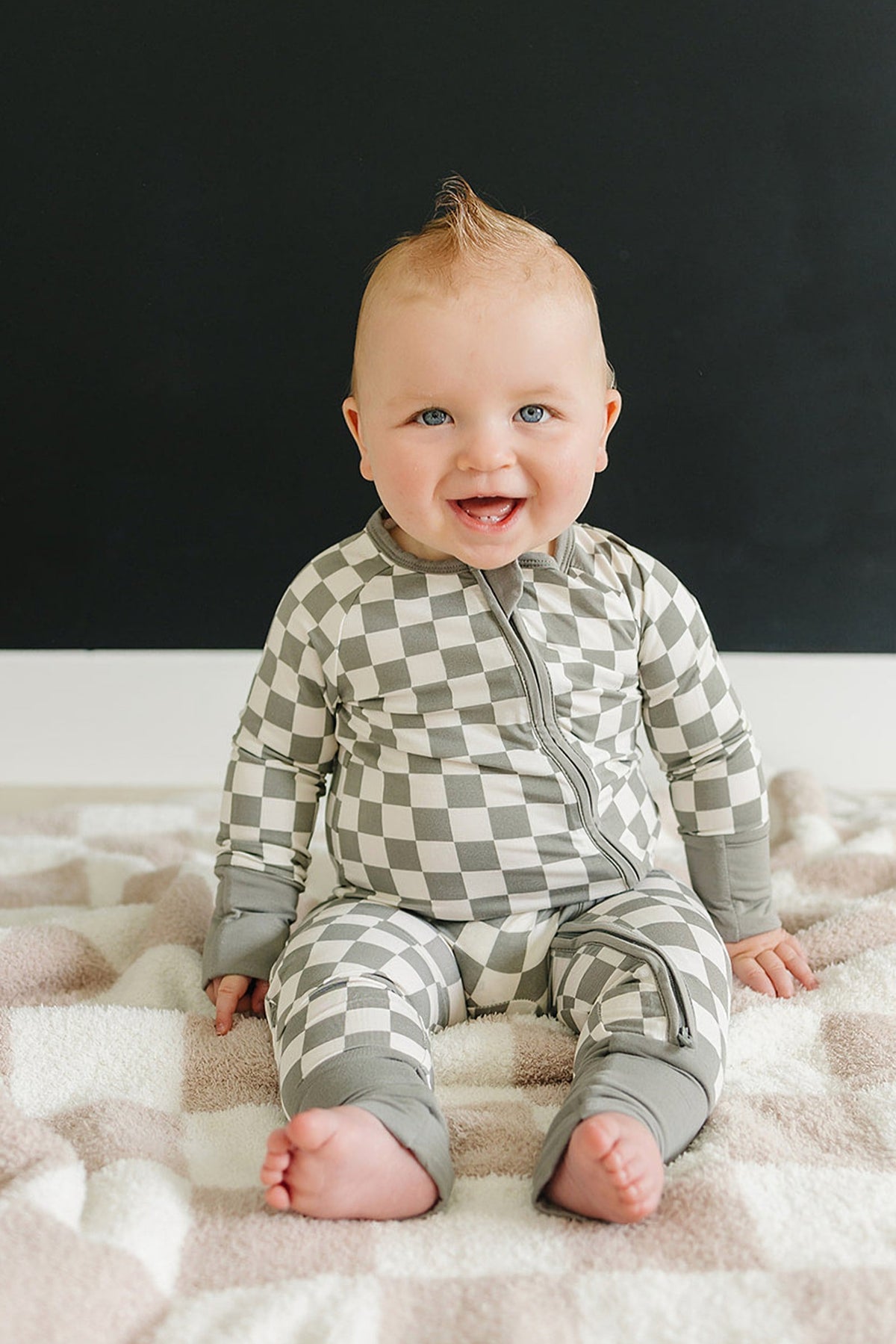 A smiling baby wears light green checkered pajamas with a zipper, sitting on a soft patterned blanket.