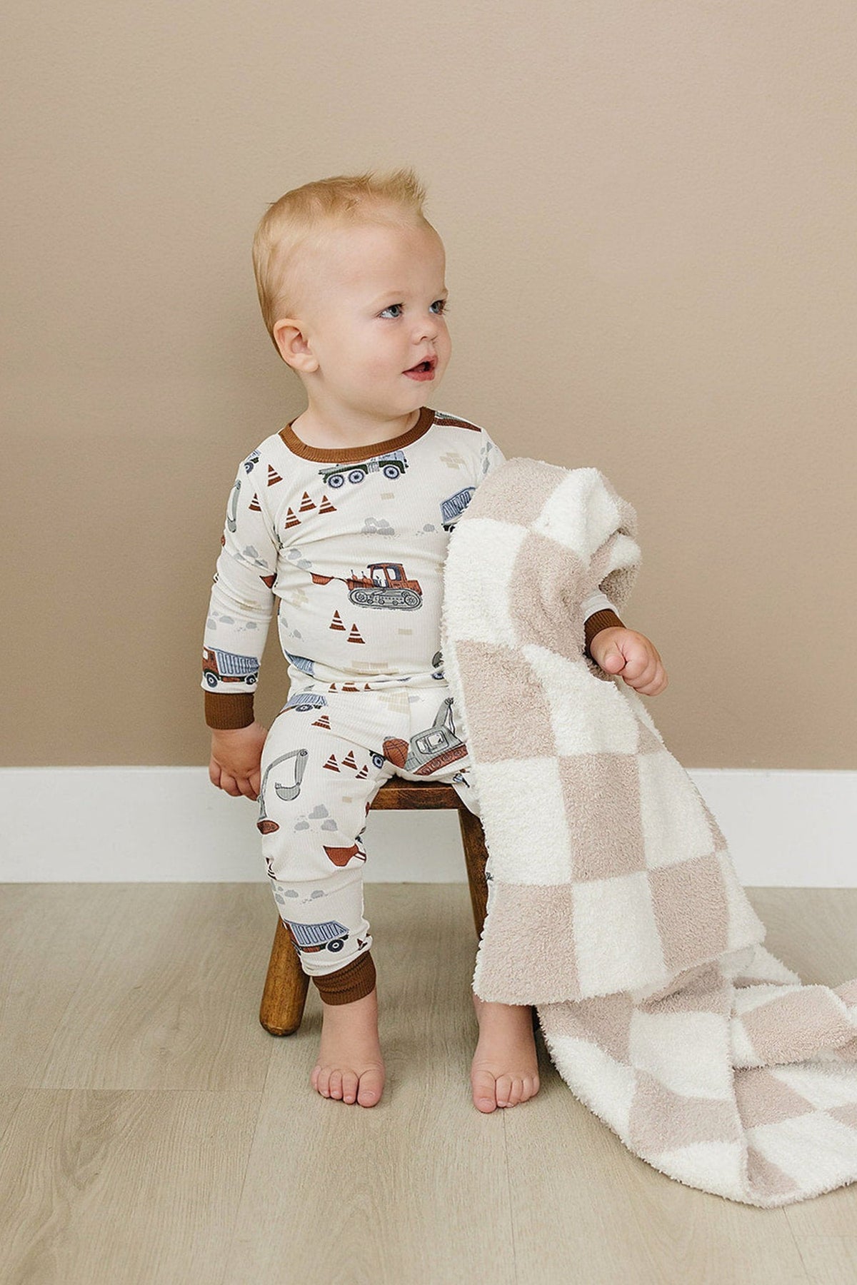 A toddler sits on a wooden stool, wearing a cozy pajama set with a construction truck pattern. The soft bamboo fabric is complemented by a plush, checkered blanket.