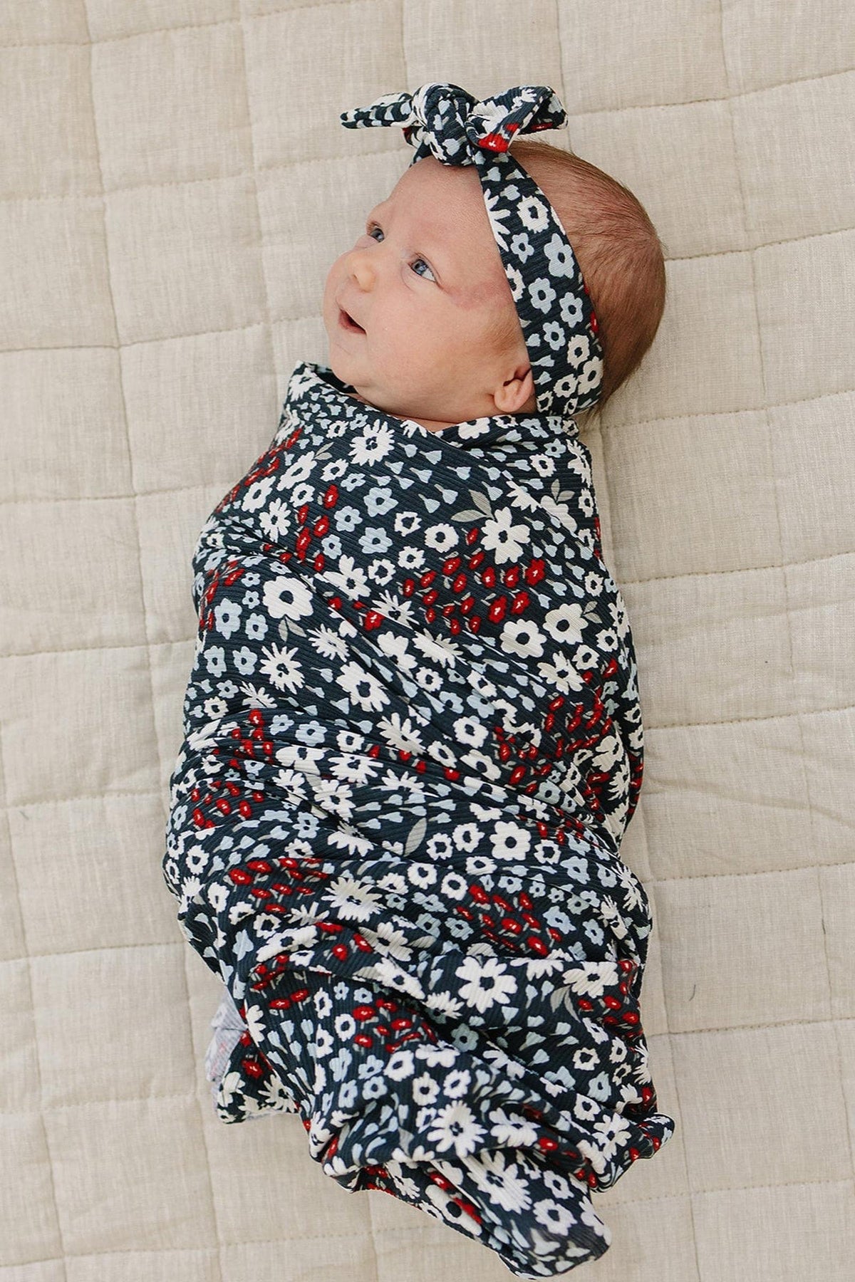 A baby wrapped in a navy floral swaddle with a matching head wrap, lying on a beige quilted surface.